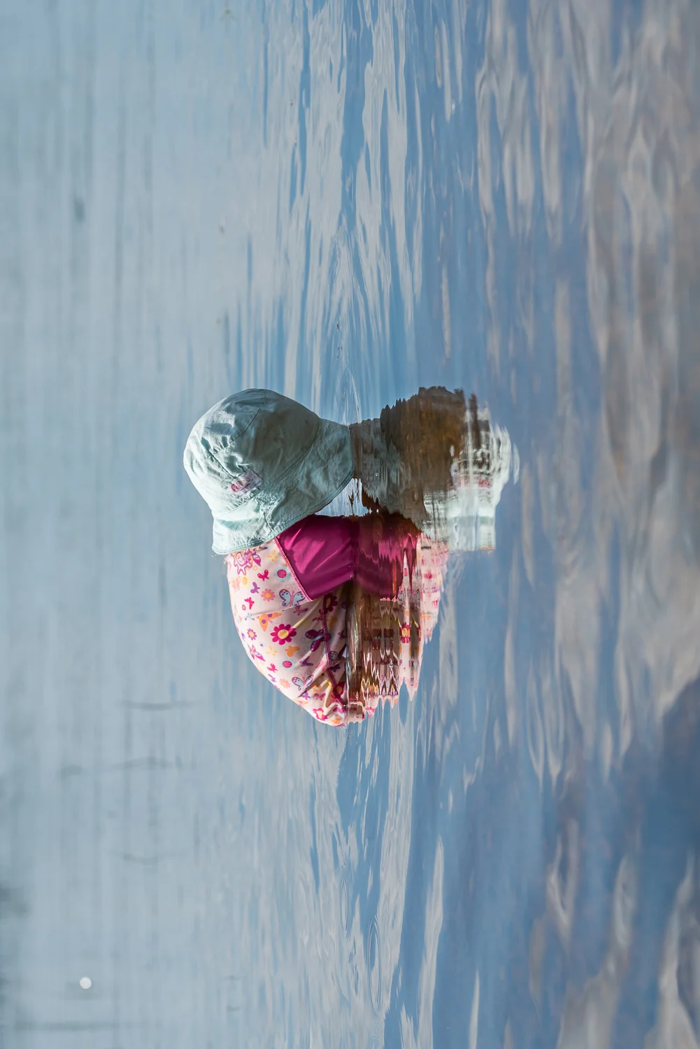 Toddler in a blue sun hat and a pink UV shirt with butterflies and flowers with her face in the blue lake water and her reflection which looks like a heart.