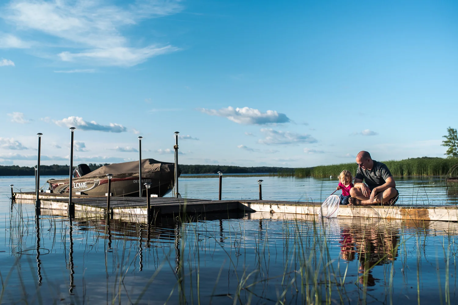 Toddler girl and her dad sitting on a wooden dock in a lake with a boat and a fishing net trying to catch treasures in the lake. Blue Sky and fluffy white clouds reflected in the blue lake water.