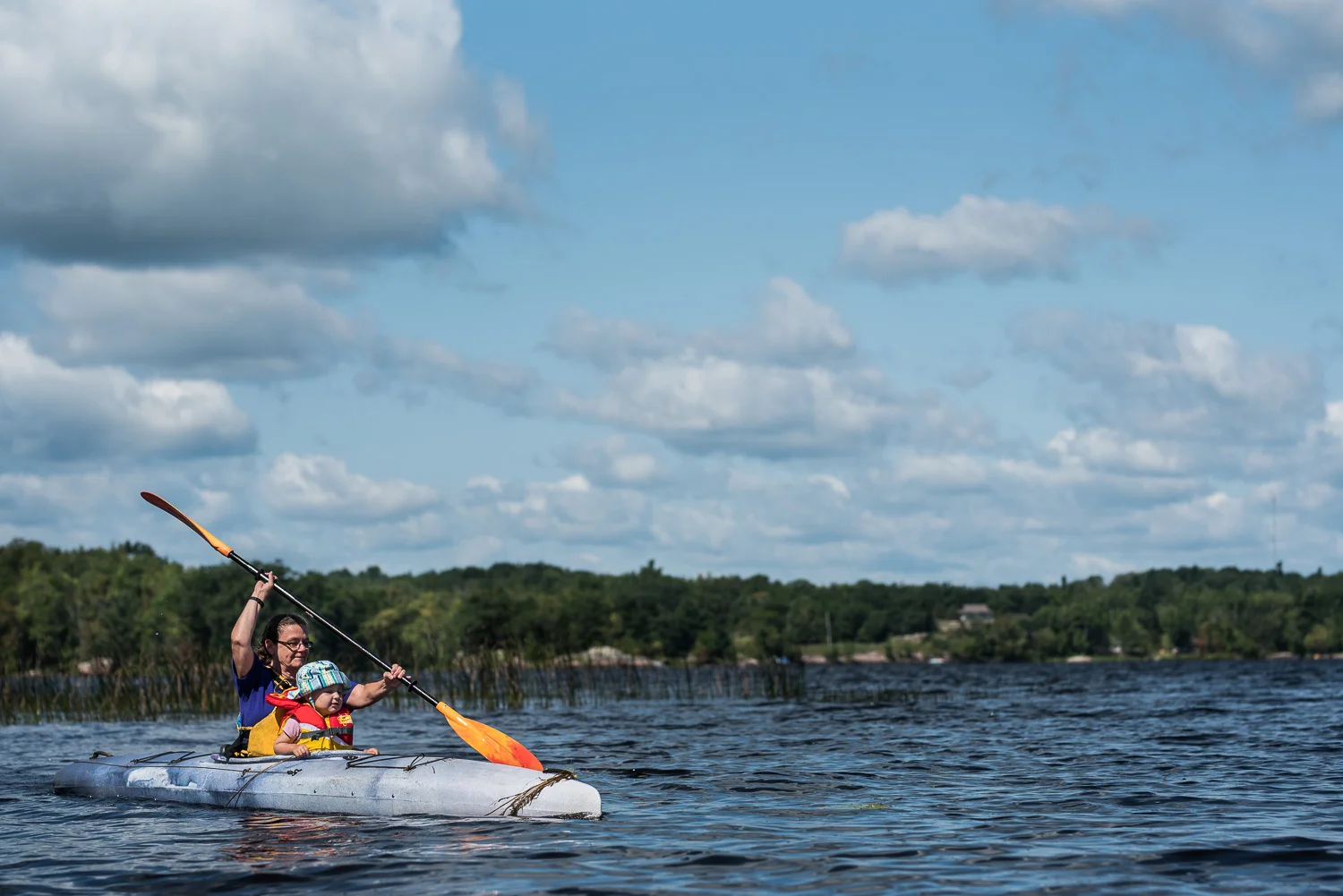 Toddler girl in blue sun hat and orange and yellow lifejacket with Grandma wearing a purple t-shirt with a yellow life jacket in a blue kayak and a yellow and orange paddle on a blue lake, surrounded by green trees on the horizon and a blue sky with white fluffy clouds.