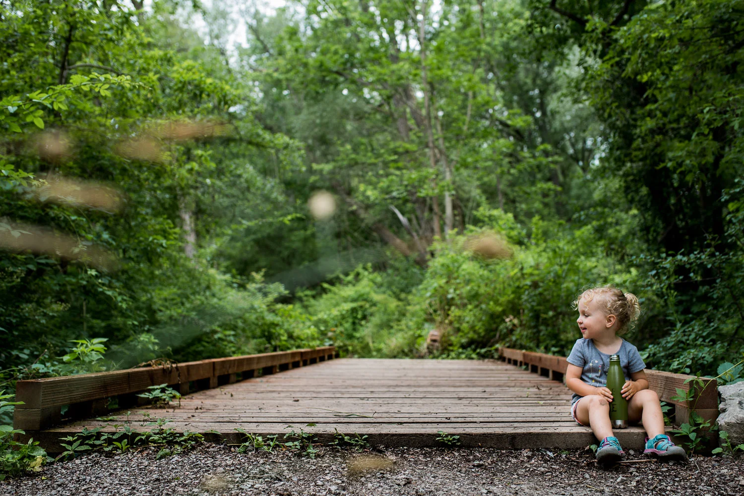 Toddler girl in a grey shirt and light blue sandals sitting on a wooden bridge surrounded by green trees and a grey gravel trail.