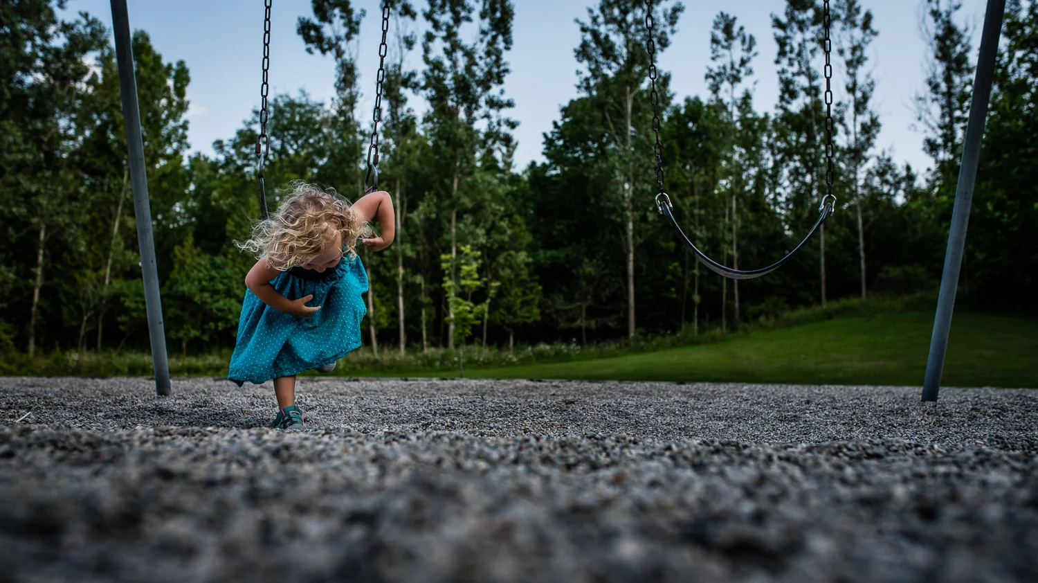 Toddler girl with curly hair in a teal blue dress laying on her tummy on a big kid swing over grey gravel with green trees and a blue sky in the background