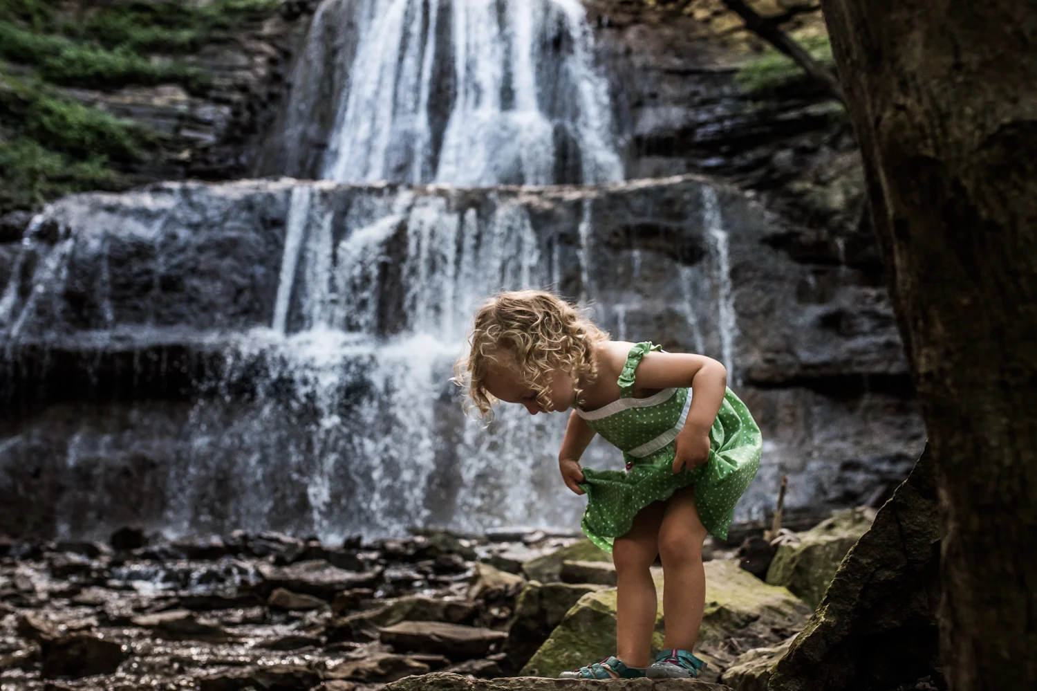 Toddler girl with blond curly hair in a green and white polka-dot dress holding up her skirt while standing on a rock with at Sherman Falls trail in Ancaster with a waterfall in the background