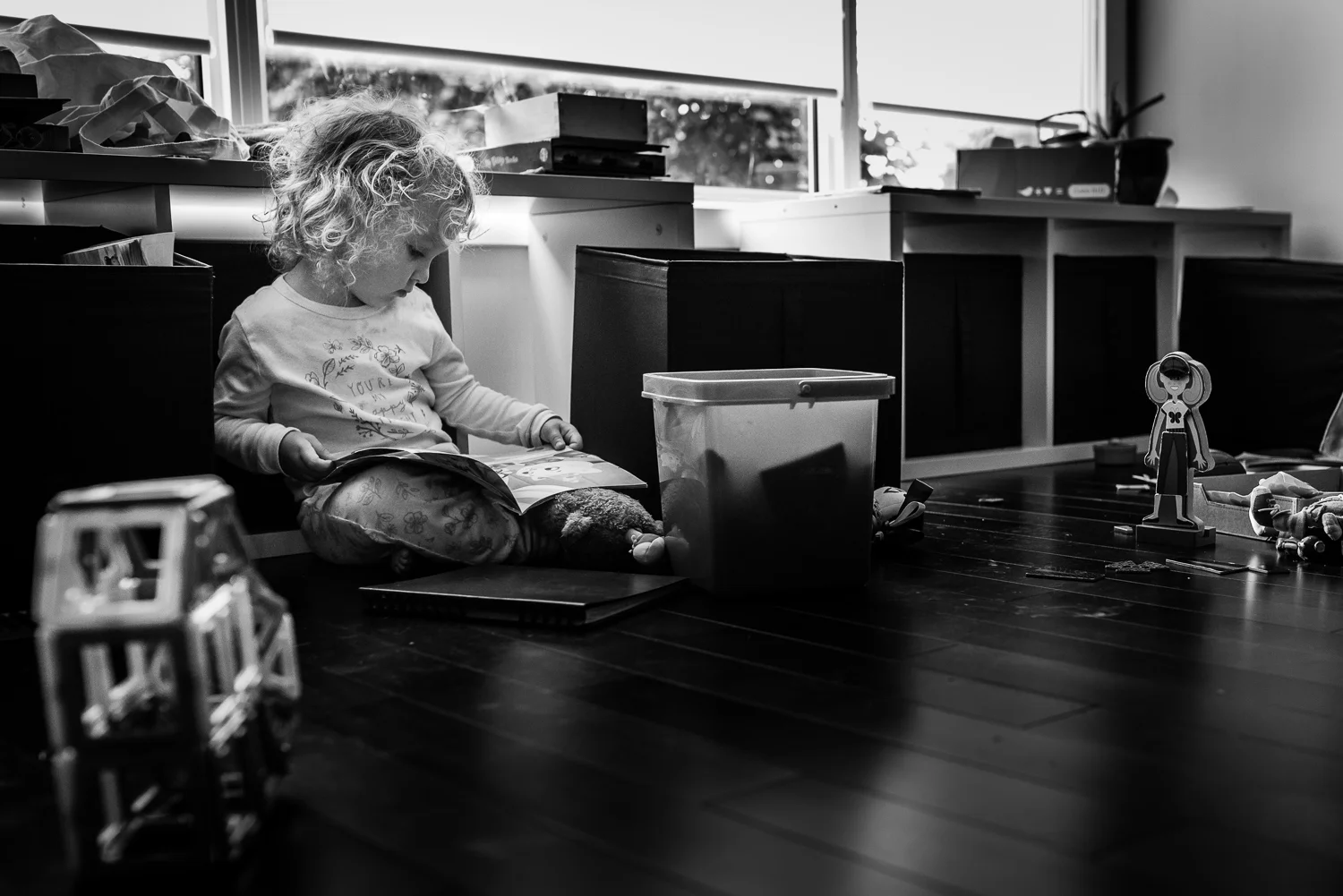 Black and white photograph of a toddler girl with curly hair sitting on the floor surrounded by toys and reading a book