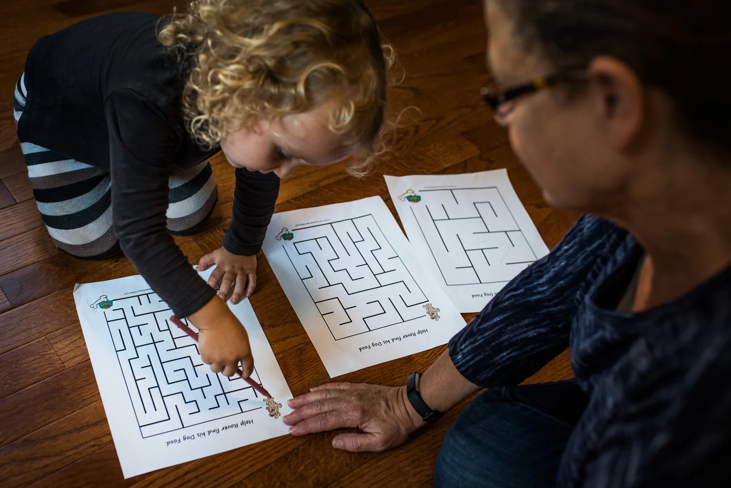 Toddler girl with curly hair sitting on the floor with her grandma working on mazes printed out on white paper
