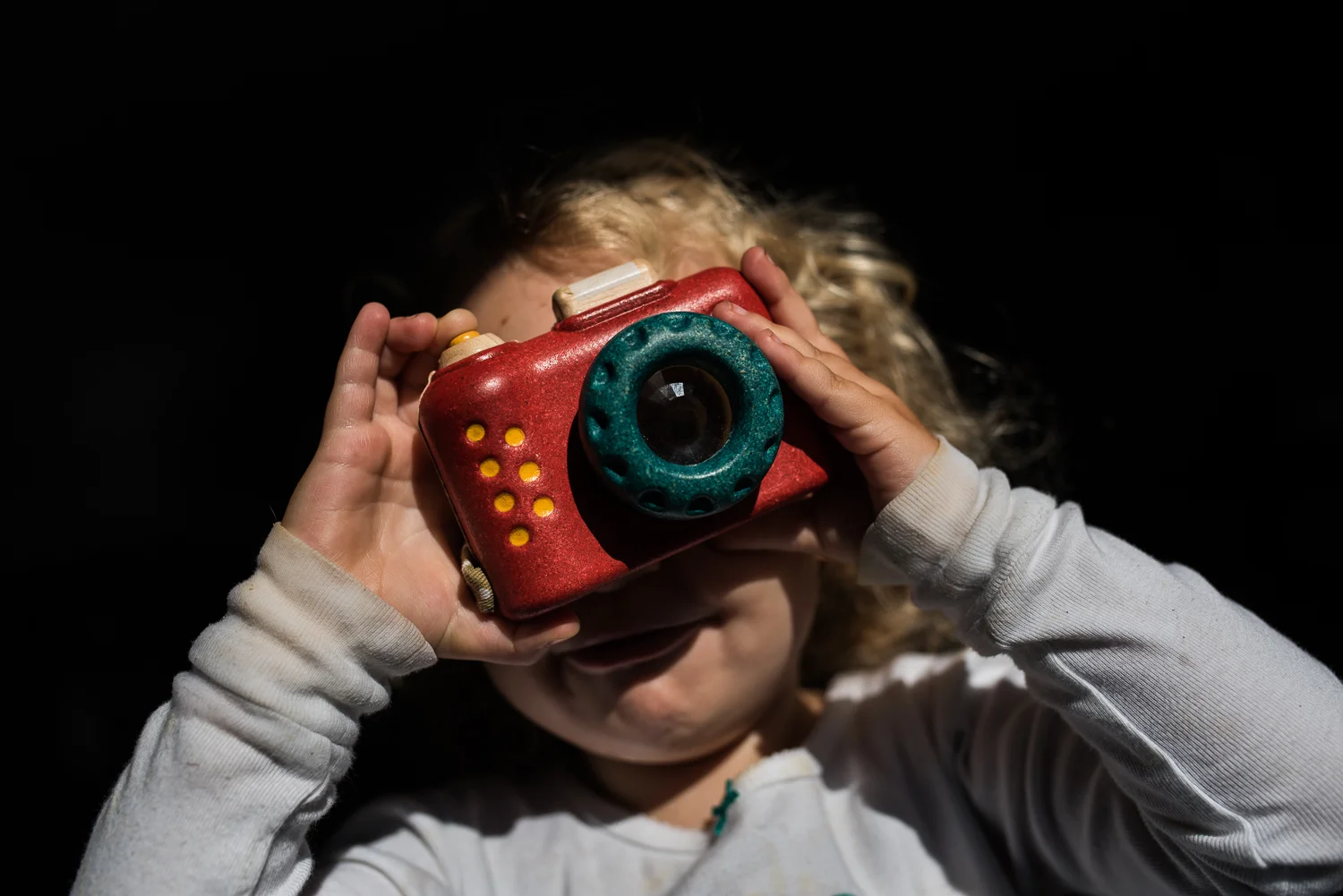 A toddler girl with curly hair holding a red, yellow and green toy camera to her eye