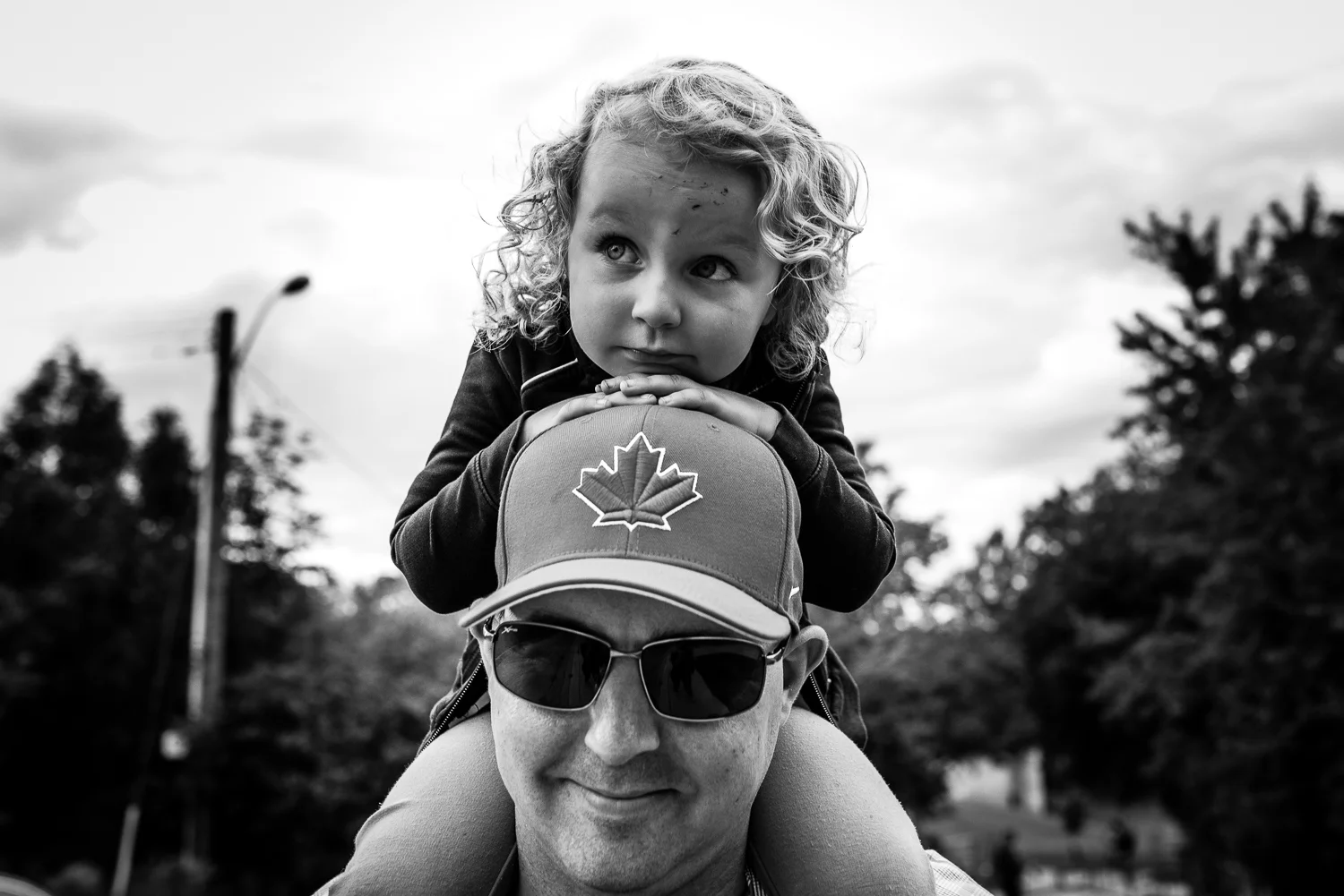 A black and white image of a toddler girl with curly hair sitting on her dad's shoulders resting her hands and head on his head and hat with a canadian maple leaf on it.