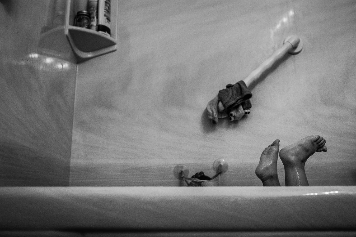 Black and white image of a bathtub with a few toys scattered about and two small toddler feet sticking out without being able to see who they are attached to.