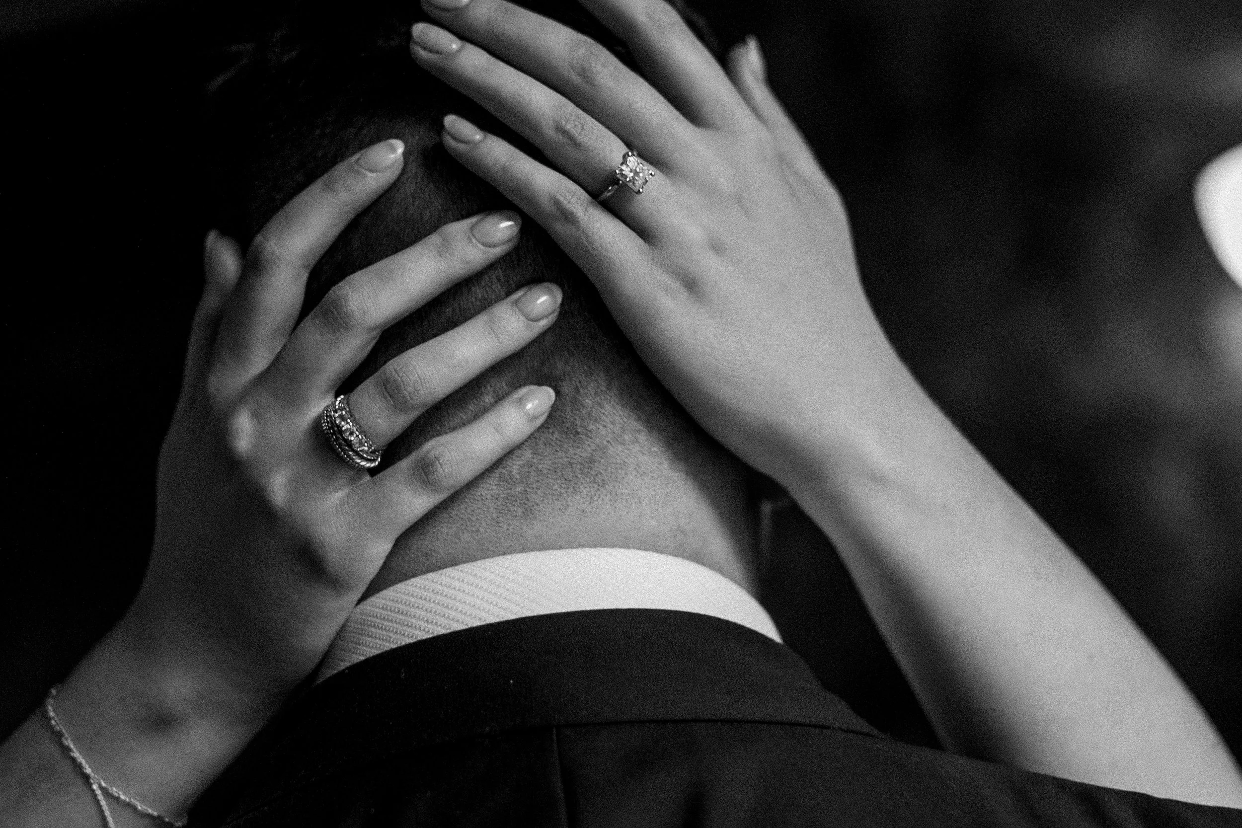 Close-up of a couple embracing, with the woman's hands on the man's head, showcasing rings on her fingers, in black and white.