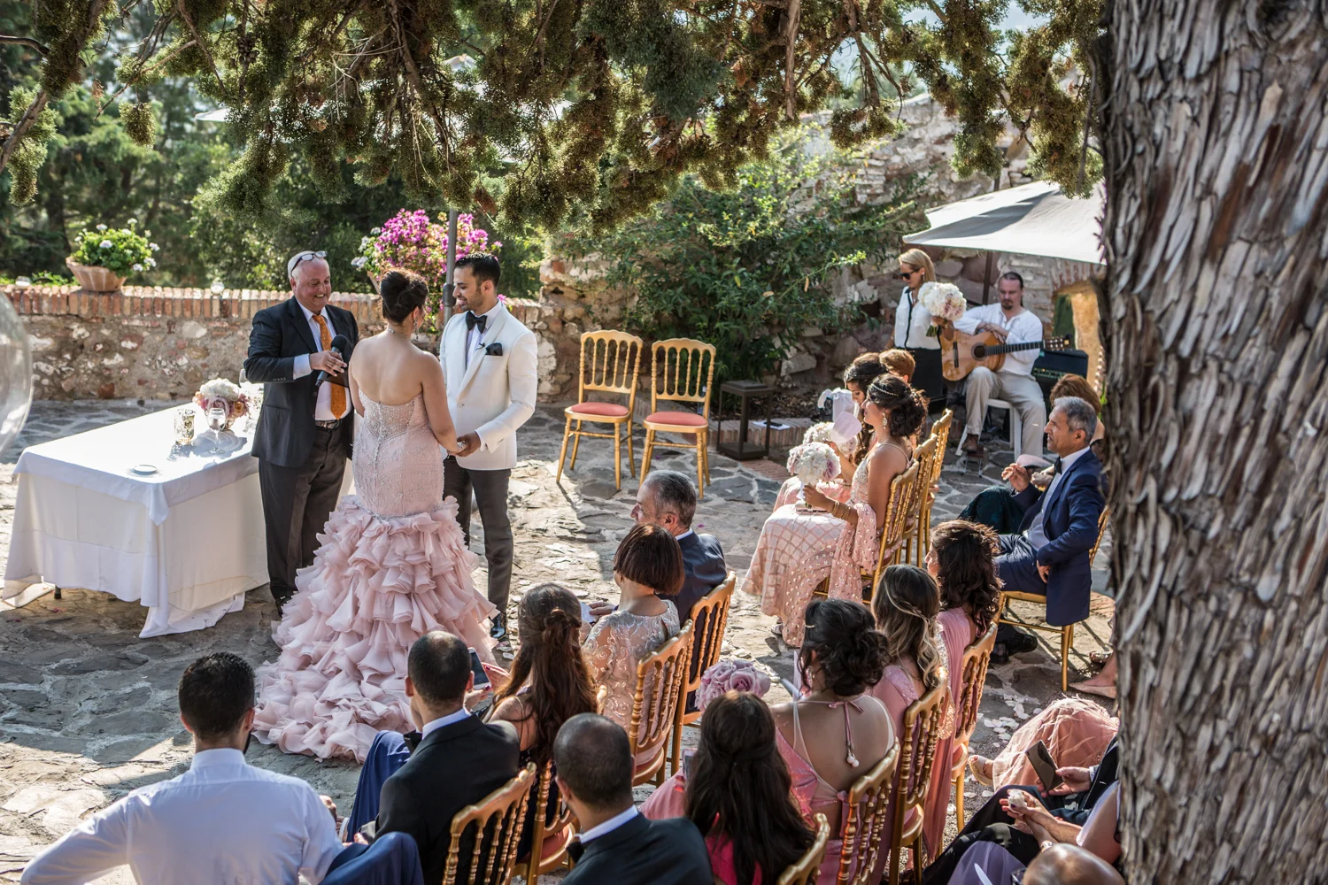 An outdoor wedding ceremony with a bride, groom, officiant, and guests seated on gold chairs among trees and plants. The officiant is holding a microphone, and the groom is dressed in a white suit with a black bow tie. Bridesmaids hold bouquets, and 