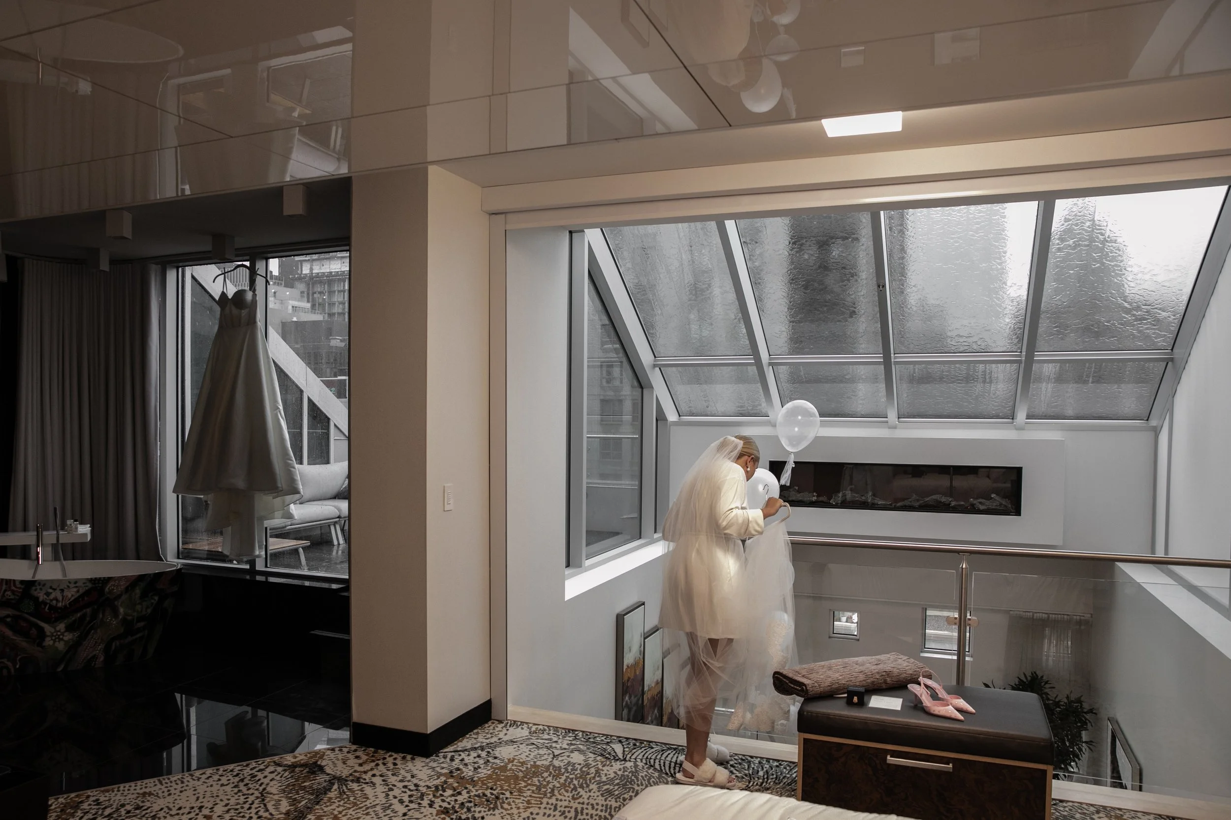 A woman wearing a white dress and veil standing near a large glass window or skylight, holding a balloon and looking at her phone in a modern, well-lit room with a fireplace and decorative elements.