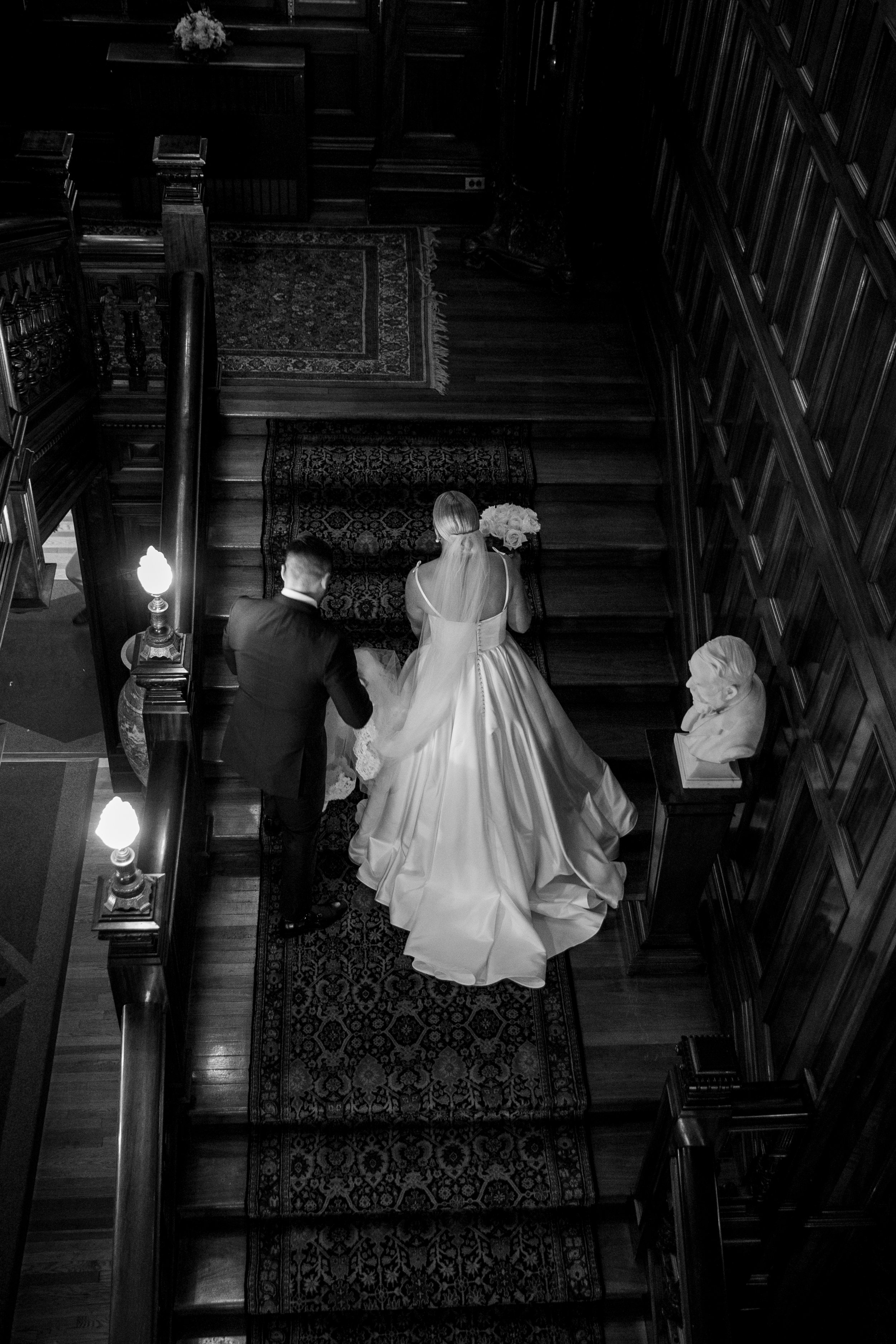 A black-and-white photo of a bride in a wedding gown holding a bouquet, walking on a staircase with a man in formal attire. The staircase has a patterned rug and wood-paneled walls, with a bust sculpture on the right.