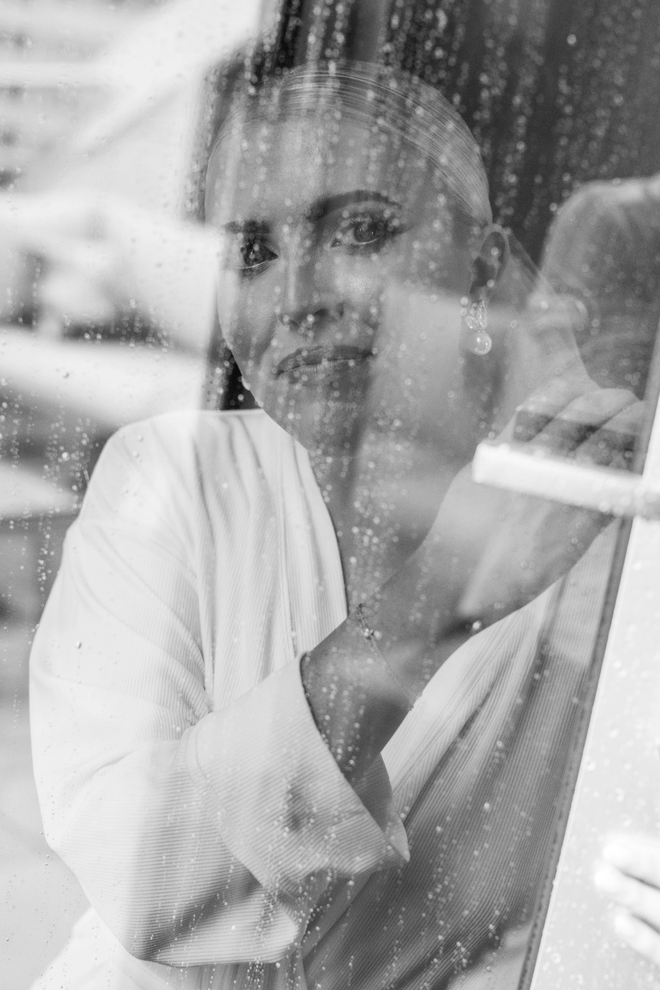 A woman with her hair slicked back, looking through a rain-covered glass window, wearing earrings and a white coat.
