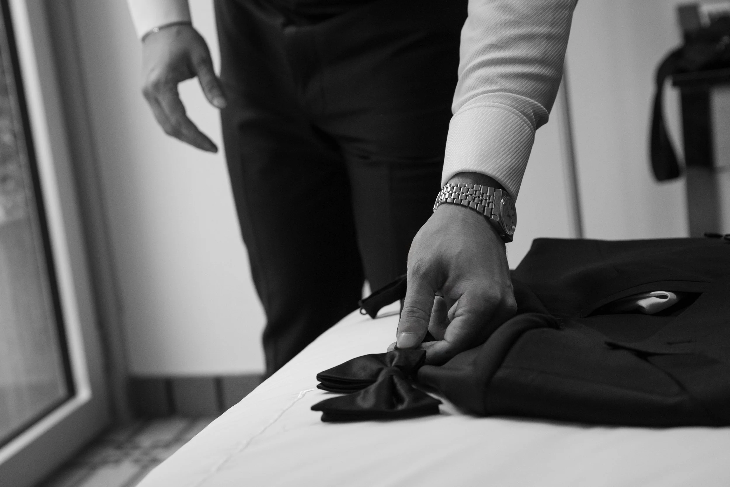 A person with a watch on their wrist is placing a tie on a black suit jacket on a bed, with a computer mouse nearby.