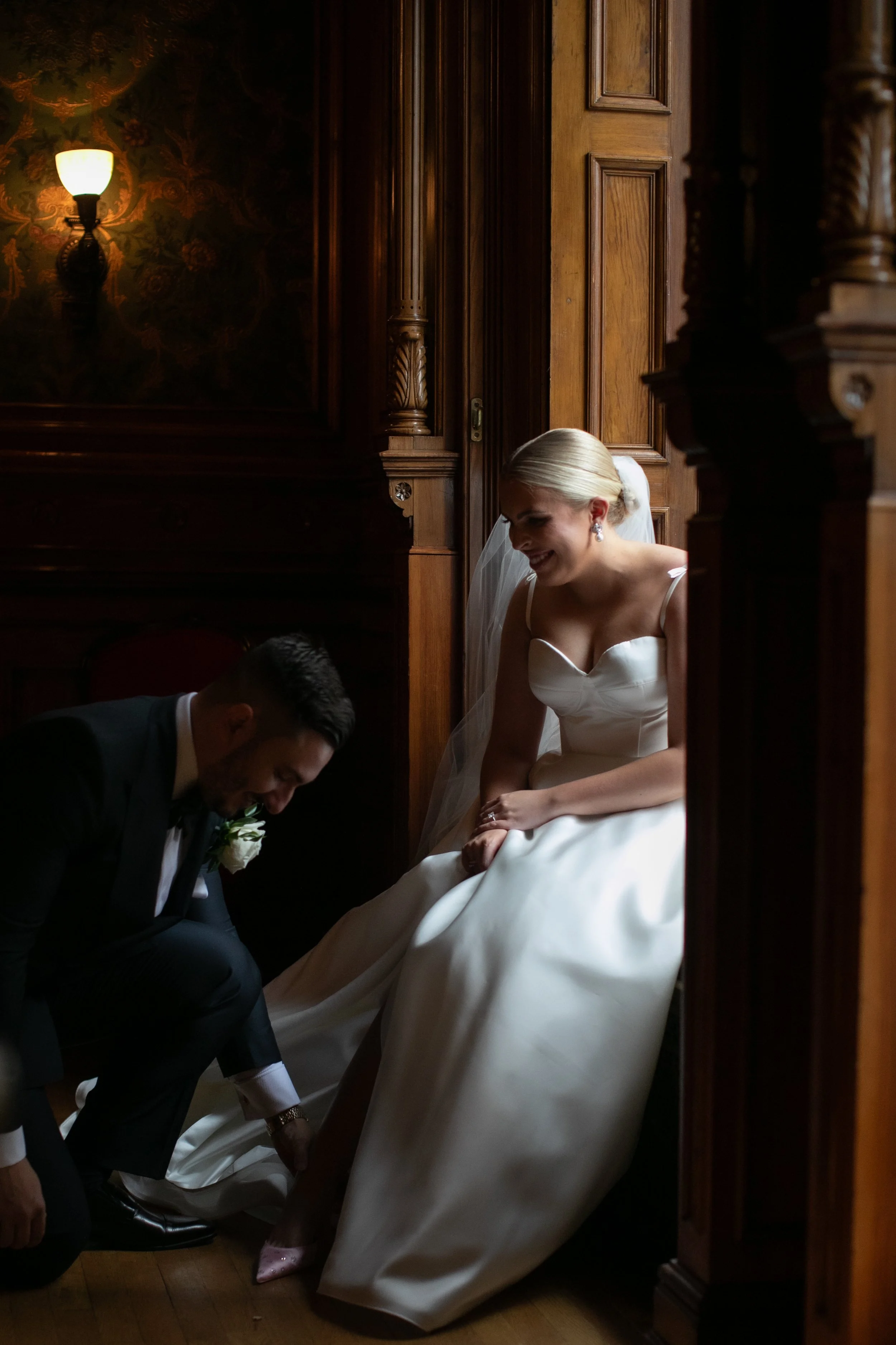A bride sitting on a wooden staircase, smiling at her groom who is kneeling and adjusting her shoe. The setting features dark wood paneling and warm lighting.