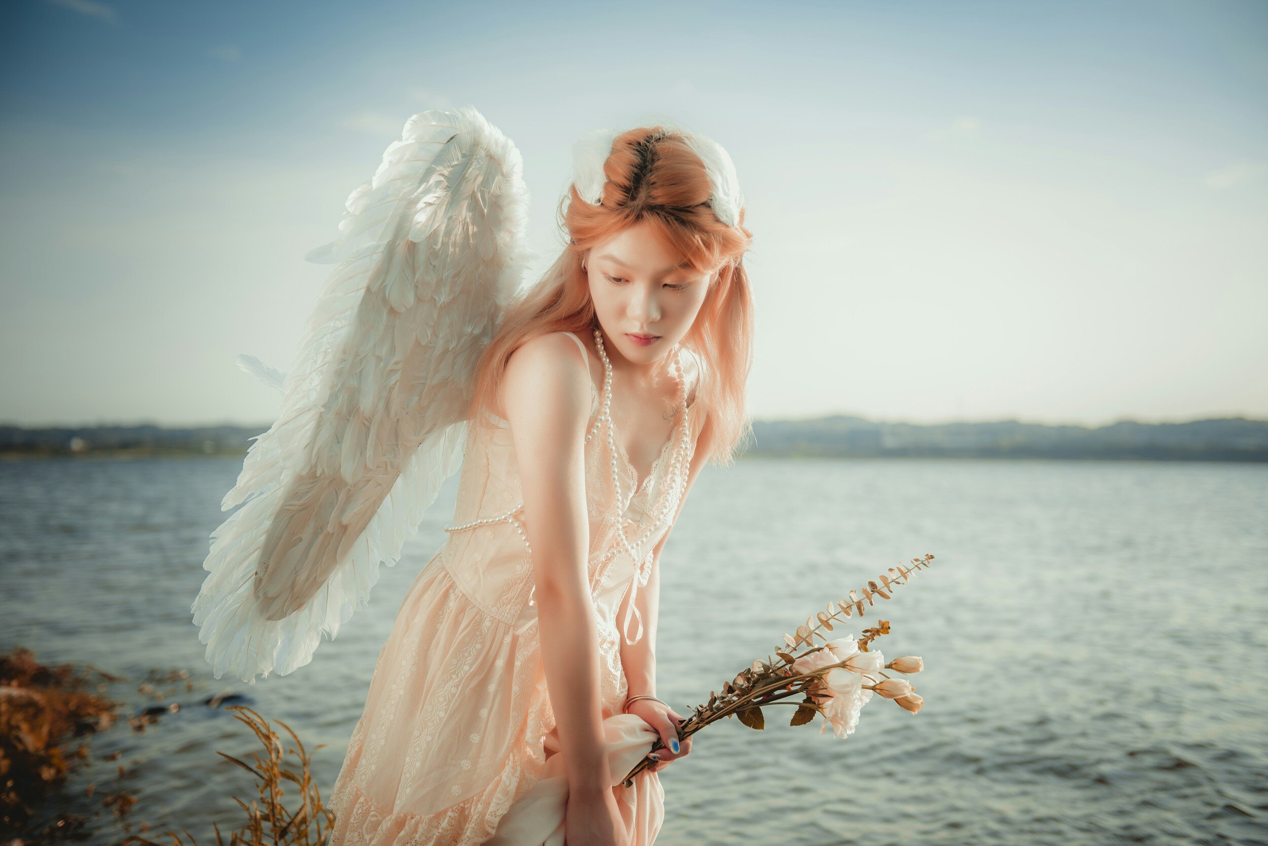 Female angel with white wings looks down while standing in front of the ocean