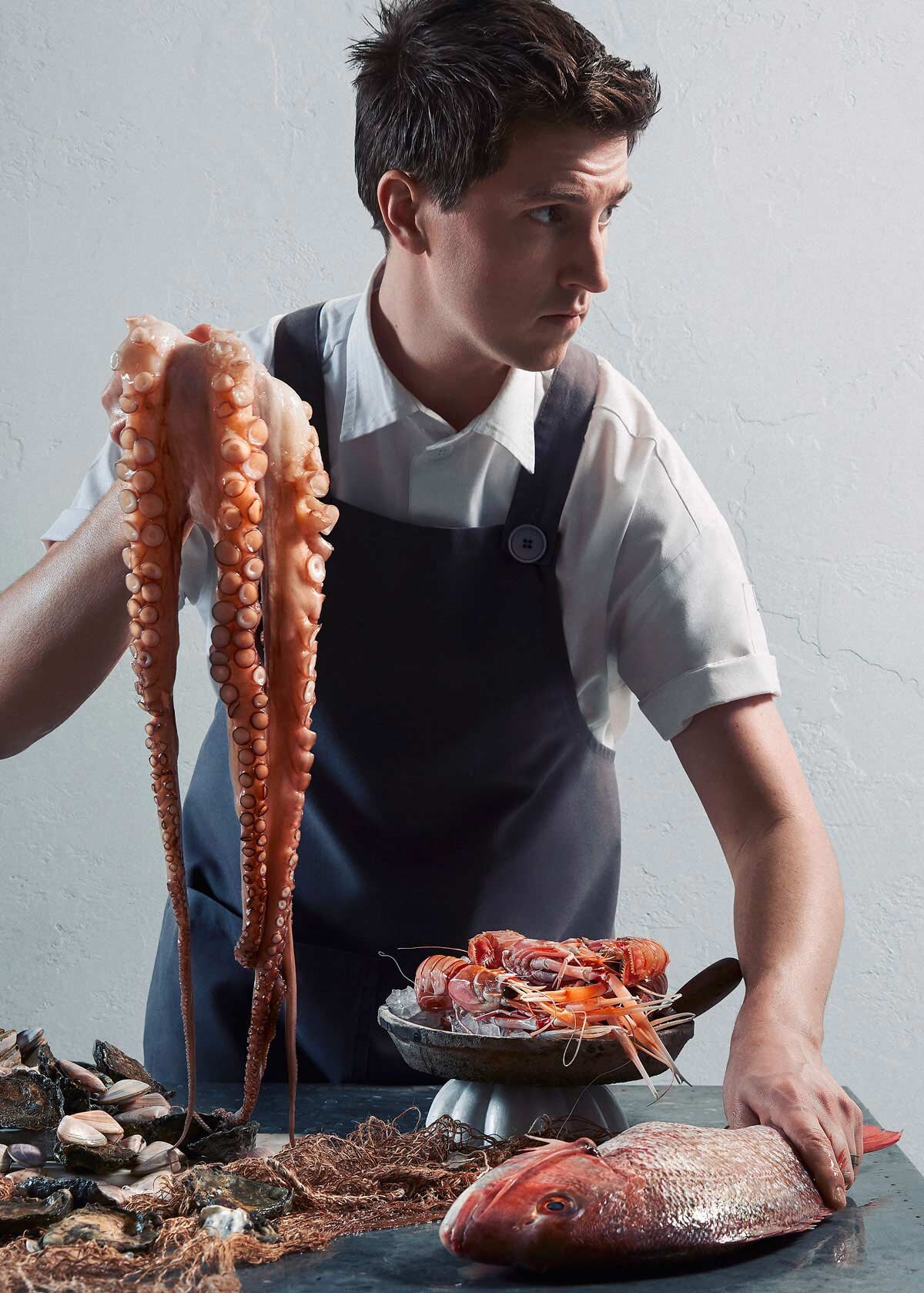  A moody portrait of Josh Niland - The talented figure behind Saint Peter and Fish Butchery holds a trailing octopus up in one hand, in front of a spead of seafood. Captured for the cover of Harvey Norman’s Gourmet Institute 2019. Art Directed by Ale
