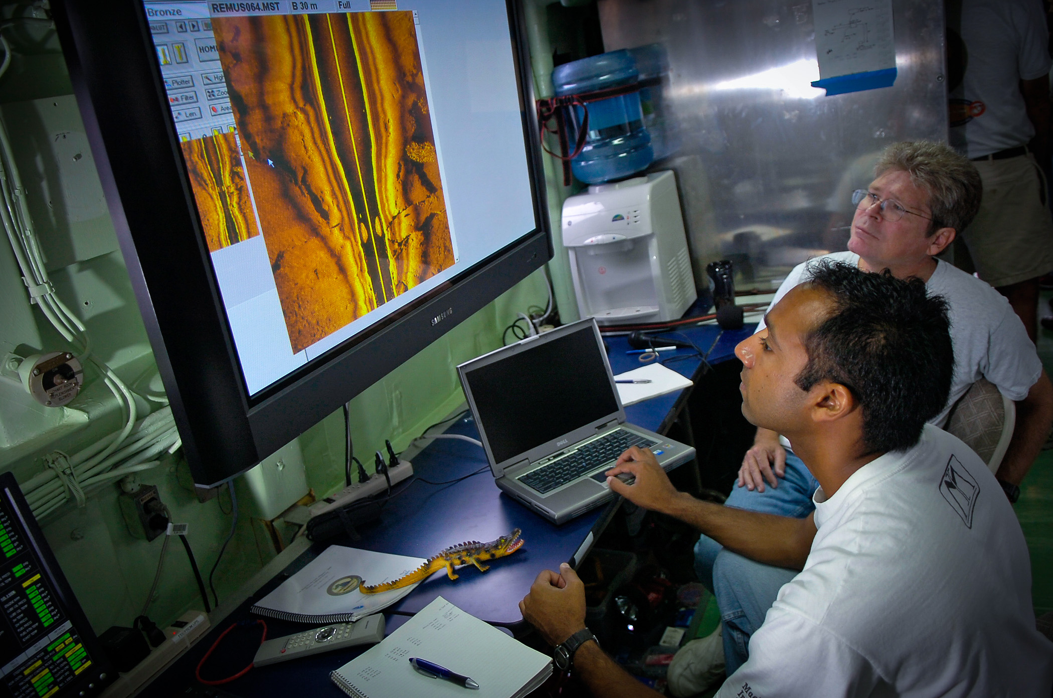 OCRACOKE, N.C. - Aamir Qaiyumi, foreground, and Mark Connelly, with Naval Sea Systems Command in Panama City, Fla., analyze data from Remote Environmental Monitoring Units (REMUS) aboard the Office of Naval Research (ONR) Afloat Lab, YP-679. The side scan sonar data were collected from an autonomous underwater vehicle (AUV) during the previous day's mission when the National Oceanographic and Atmospheric Administration, with support from ONR, returned to the "Graveyard of the Atlantic" to continue the search for the Alligator, the U.S. Navy's first submarine. The joint expedition is taking place off Cape Hatteras, N.C., where the Civil War-era vessel was lost during a fierce storm in 1863.