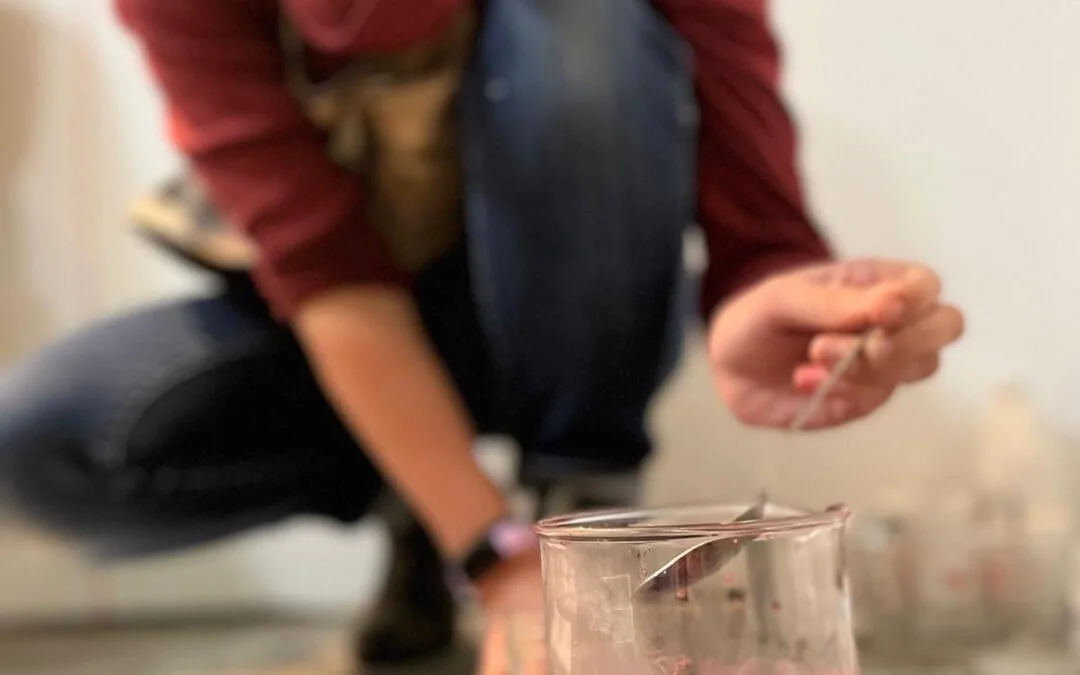 Close-up of a person's hand holding a tool above a transparent container, with the person crouching in the background.