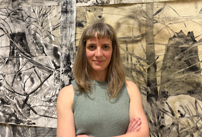 A woman with shoulder-length light brown hair standing in front of black and white abstract artwork, wearing a sleeveless green top.
