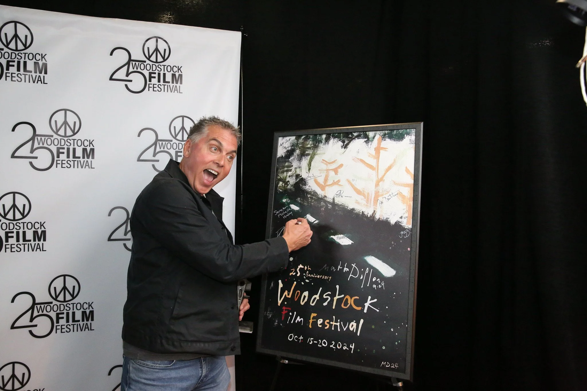A man signing a poster at the Woodstock Film Festival, October 15-20, 2024, with a backdrop displaying the festival logo.