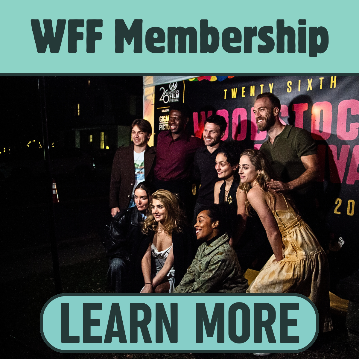 A group of people at a film event, smiling and posing for a photo in front of a backdrop with text about the Woodsstock Film Festival, with some standing and others sitting, at night.