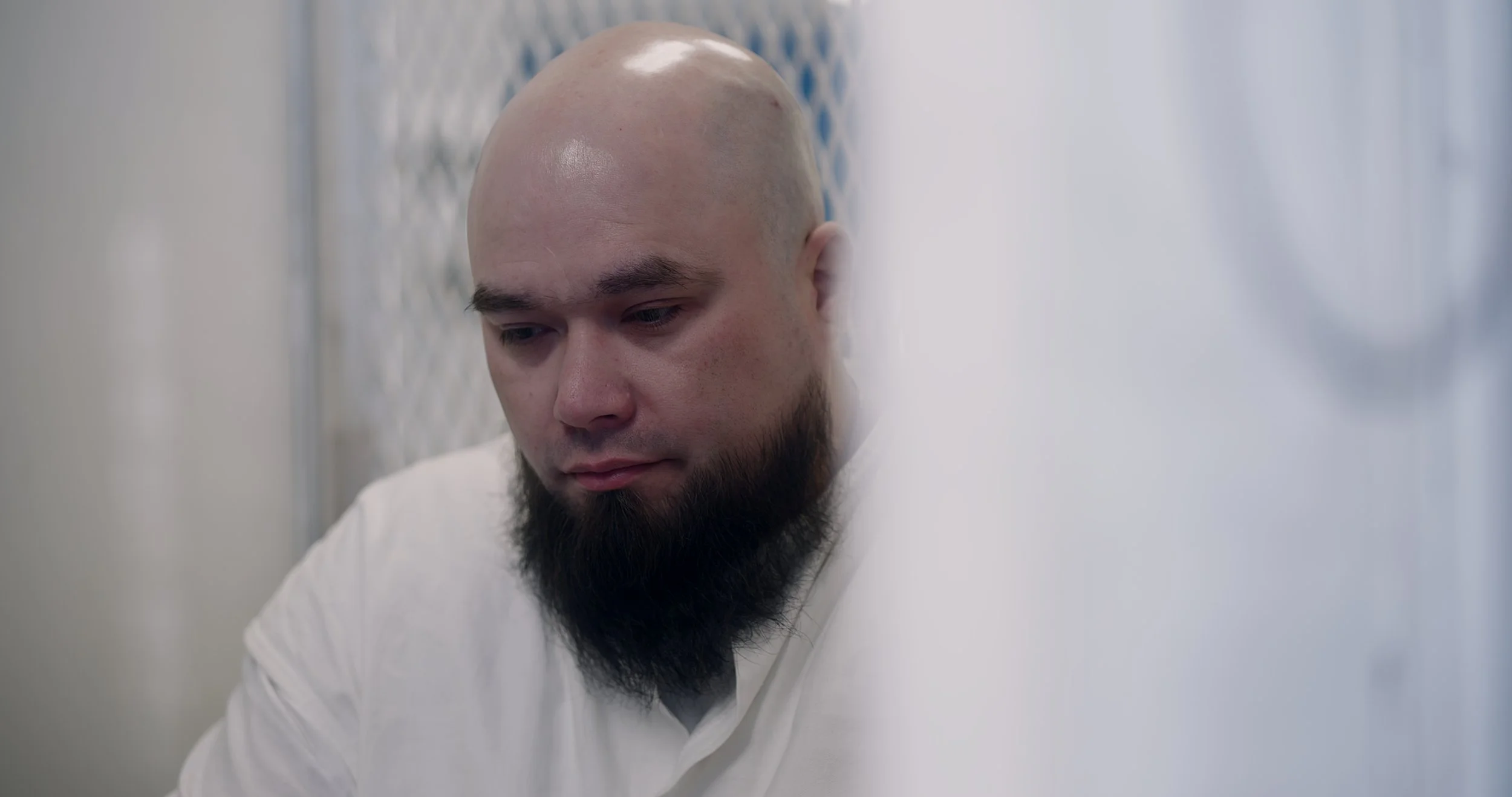 A bald man with a beard looking down in a white shirt, standing against a gray wall.