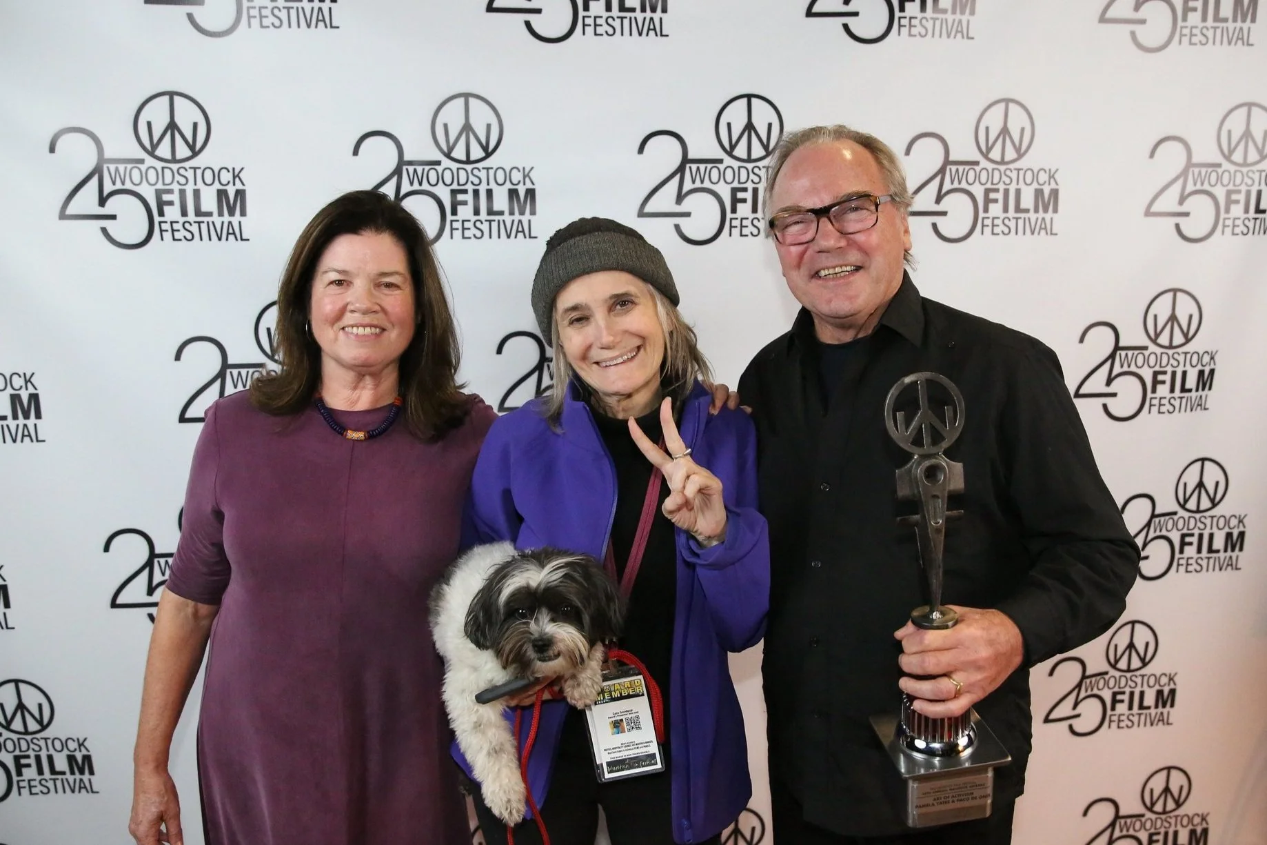Three people standing together at the 25th Woodstock Film Festival, smiling for the camera in front of a backdrop with festival logos. The woman in the middle is holding a small dog. The man on the right is holding a trophy.