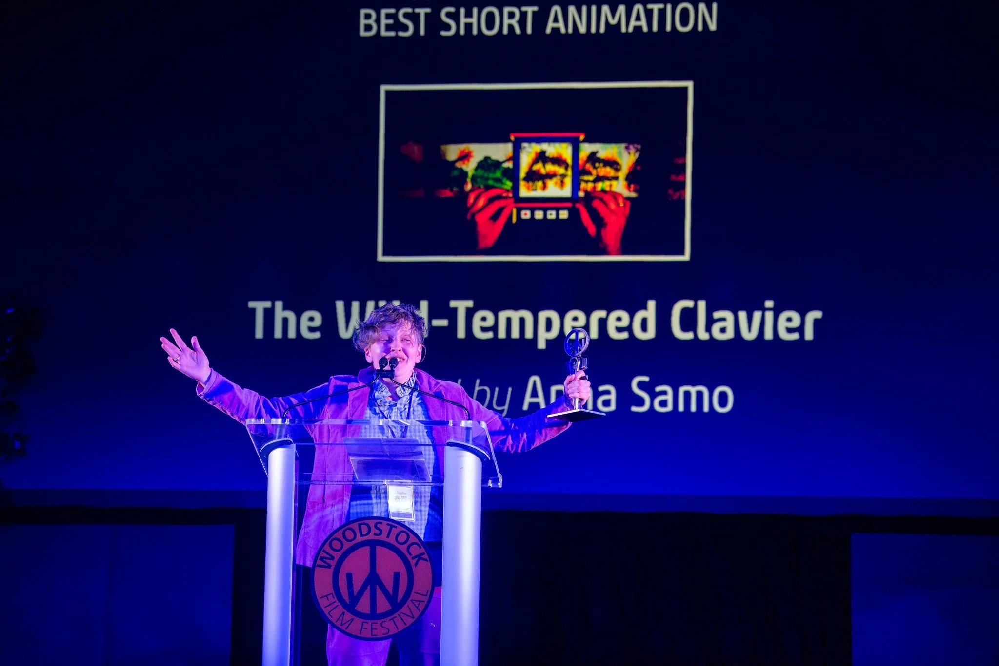 A young boy on stage at the Woodstock Film Festival holding a trophy and speaking at a podium with a certificate. Behind him is a large screen displaying an award for Best Short Animation for a film titled "The Wild-Tempered Clavier" by Anja Samo.