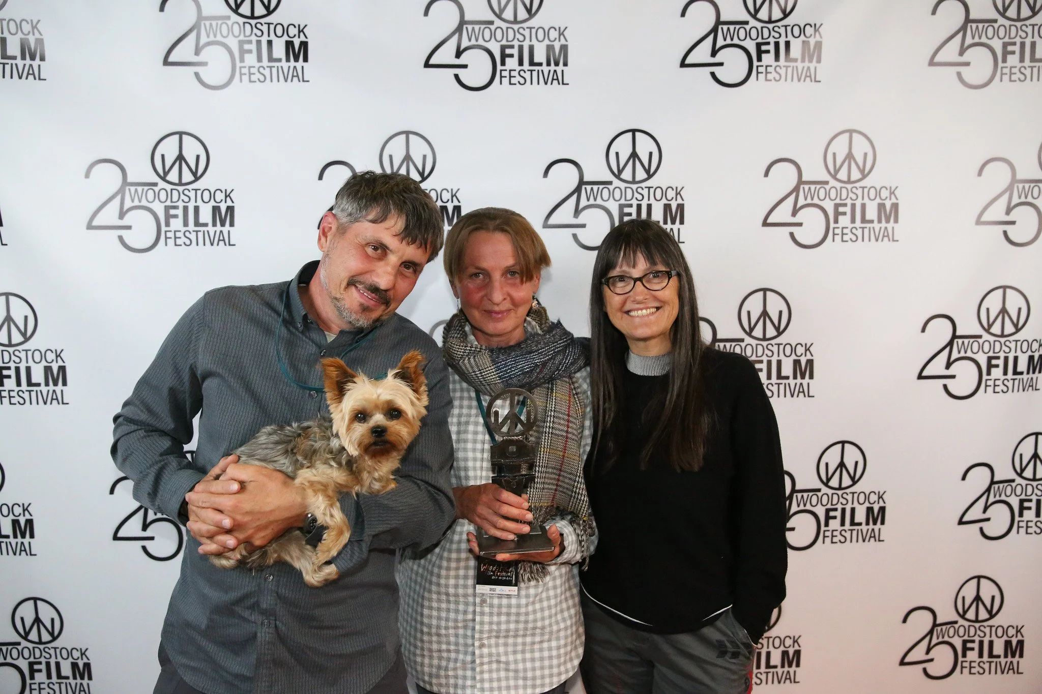 Three people standing in front of a backdrop with the words 'Woodstock Film Festival 25' and a peace symbol, smiling. The person on the left is holding a small dog. The person in the middle is holding a trophy.