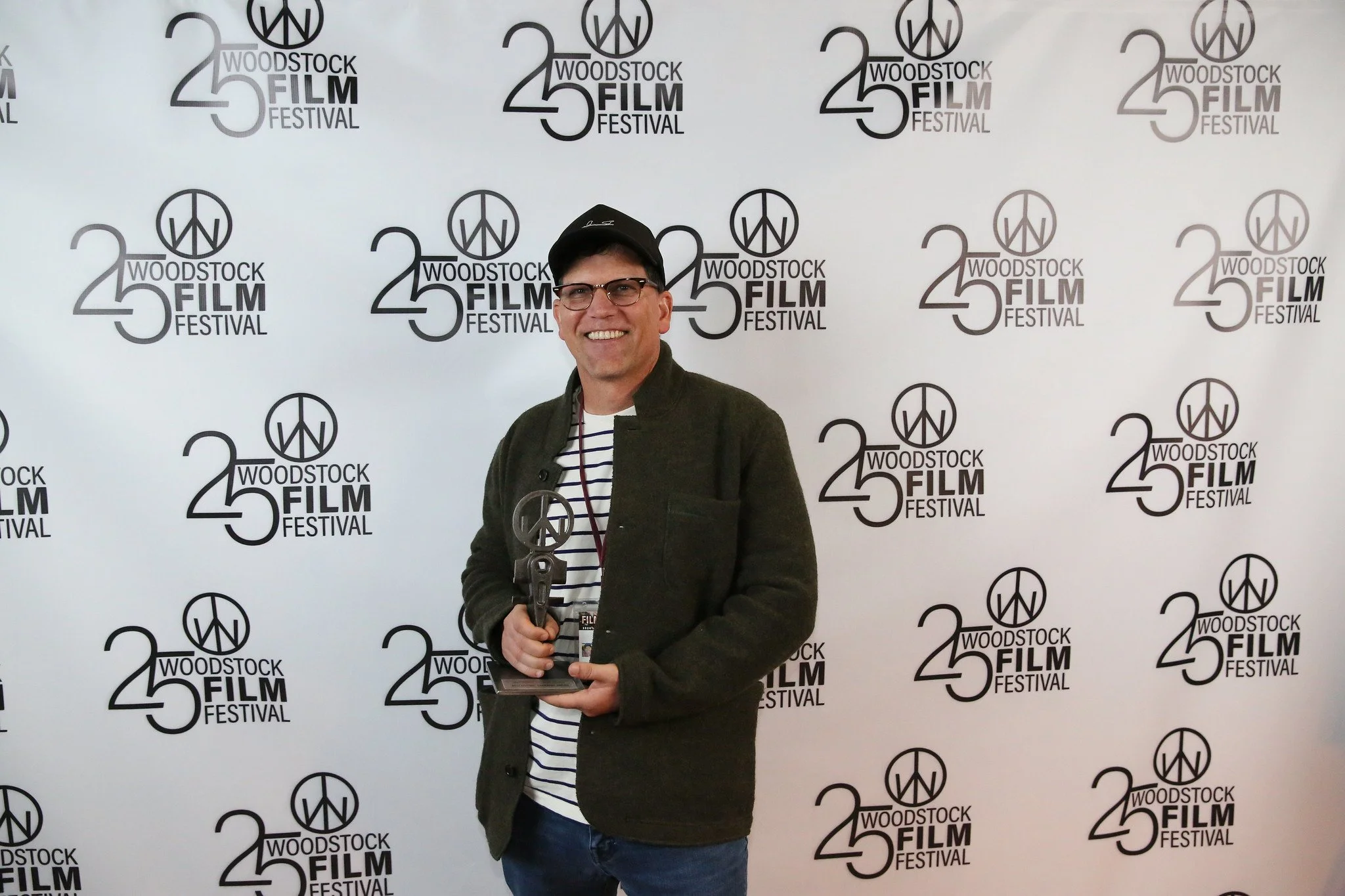 A man holding an award trophy at the Woodstock Film Festival, standing in front of a backdrop with '25 Woodstock Film Festival' logos. The man is smiling, wearing glasses, a striped shirt, a dark jacket, and a black cap.