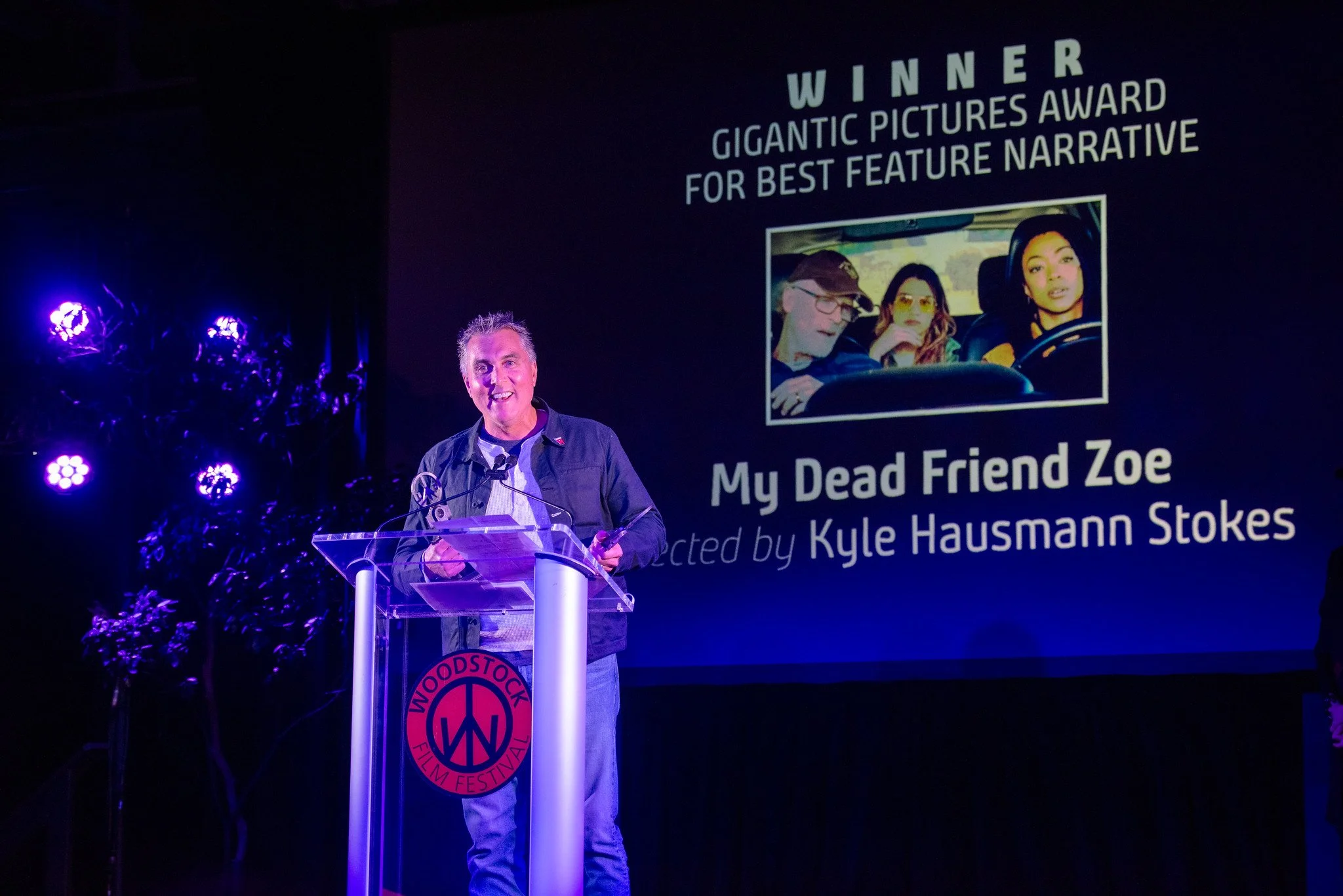 Man standing at a clear podium with a Woodstock Film Festival logo, speaking into a microphone, in front of a large screen displaying an award announcement for "My Dead Friend Zoe" directed by Kyle Hausmann Stokes, with a photo of three people in a car.