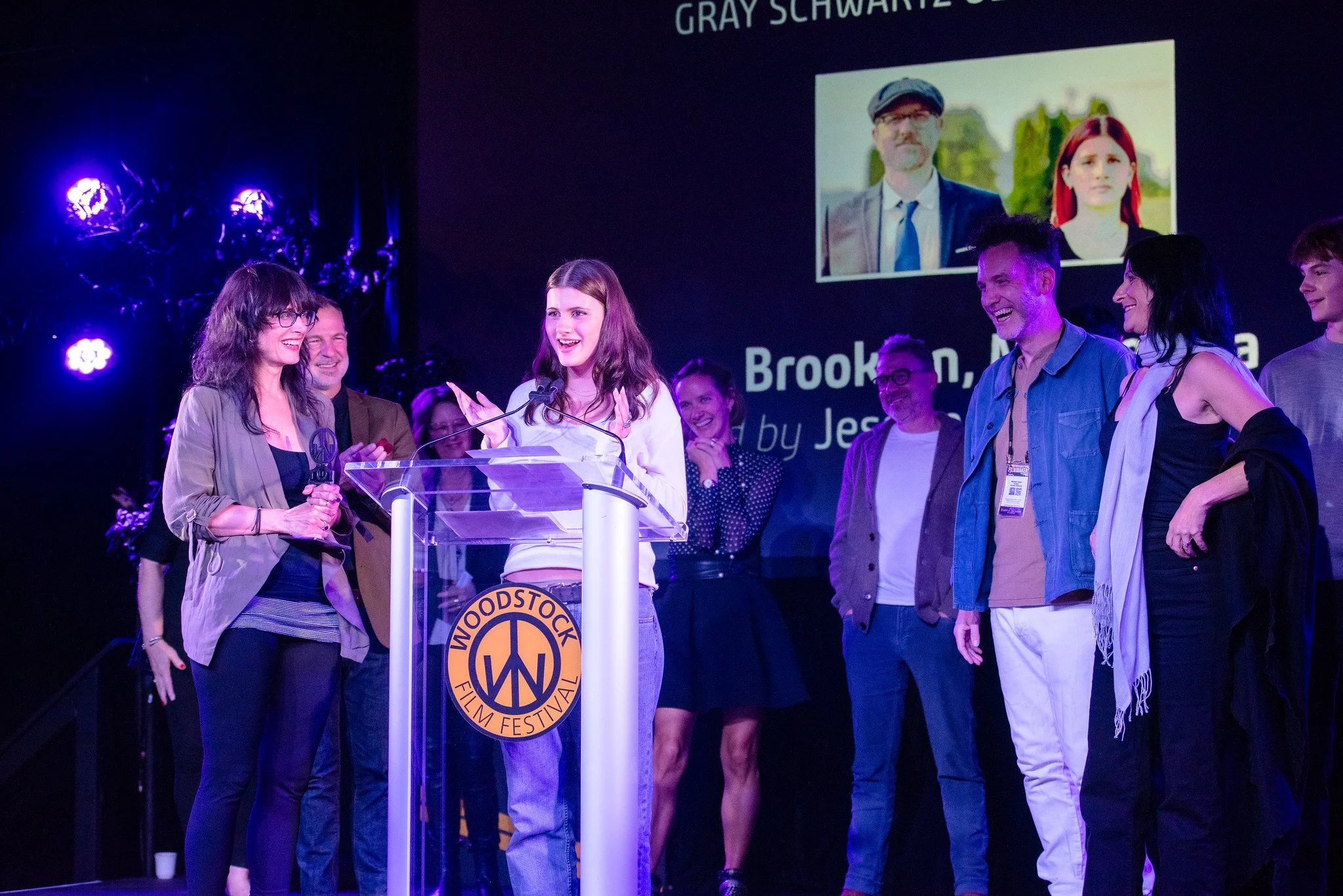 Group of people on stage at the Woodstock Film Festival, one woman speaking at a podium with the festival logo, others smiling and watching, with a large screen behind showing a picture of a man in glasses and a woman with red hair, and text including 'Brooklyn' and 'by Jessa'.