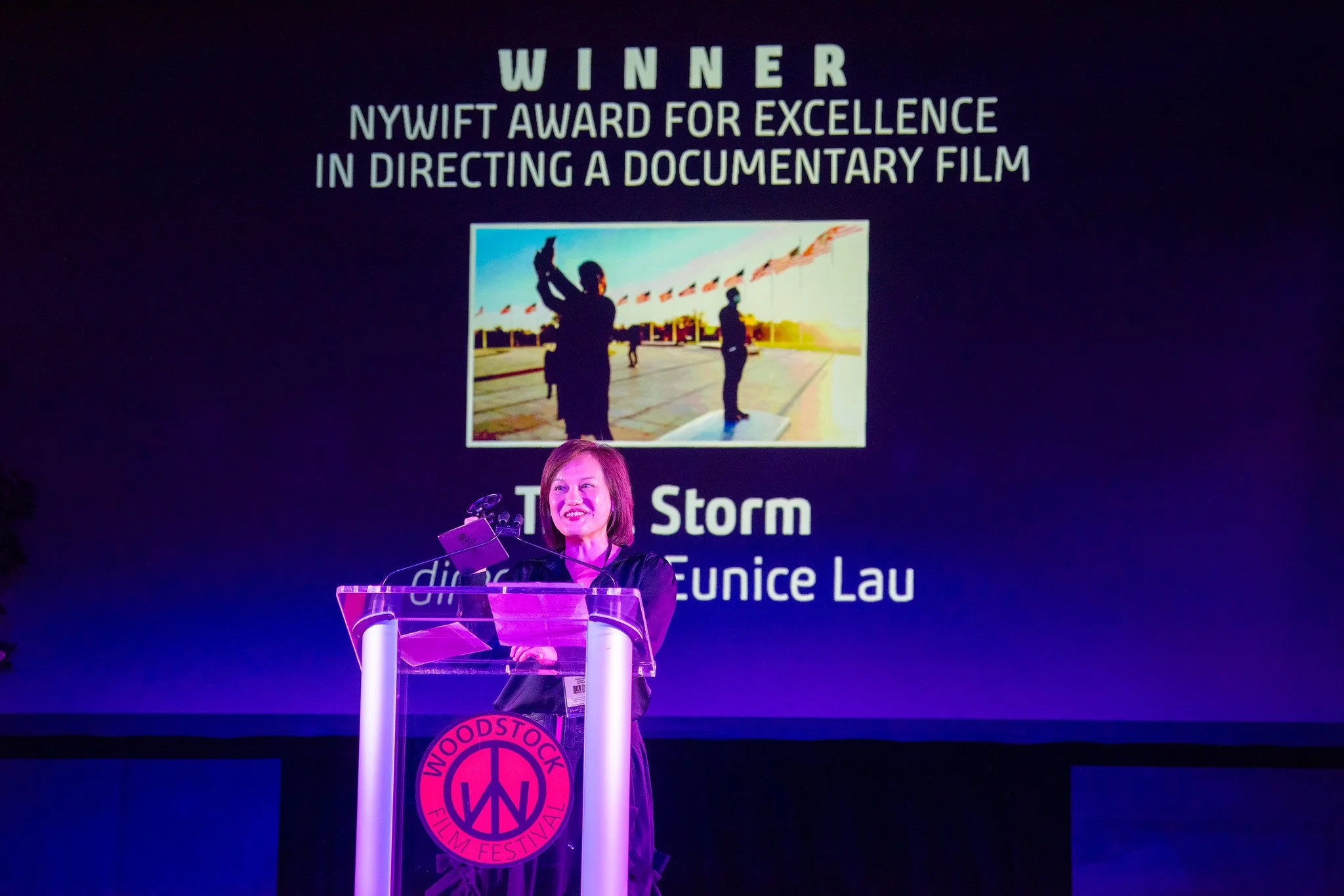 A woman stands behind a podium with a Woodstook Film Festival logo, speaking at an award ceremony, with a large screen behind her displaying her achievement of winning the NYWIFT award for directing a documentary film.