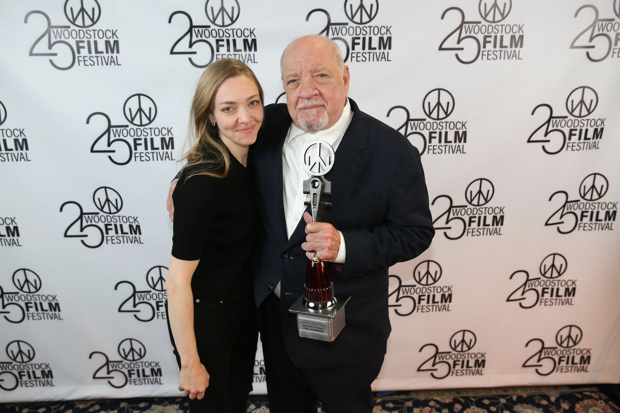 Two people, a woman and a man, standing together at the Woodstock Film Festival, holding an award trophy, with a backdrop displaying the festival's logo.