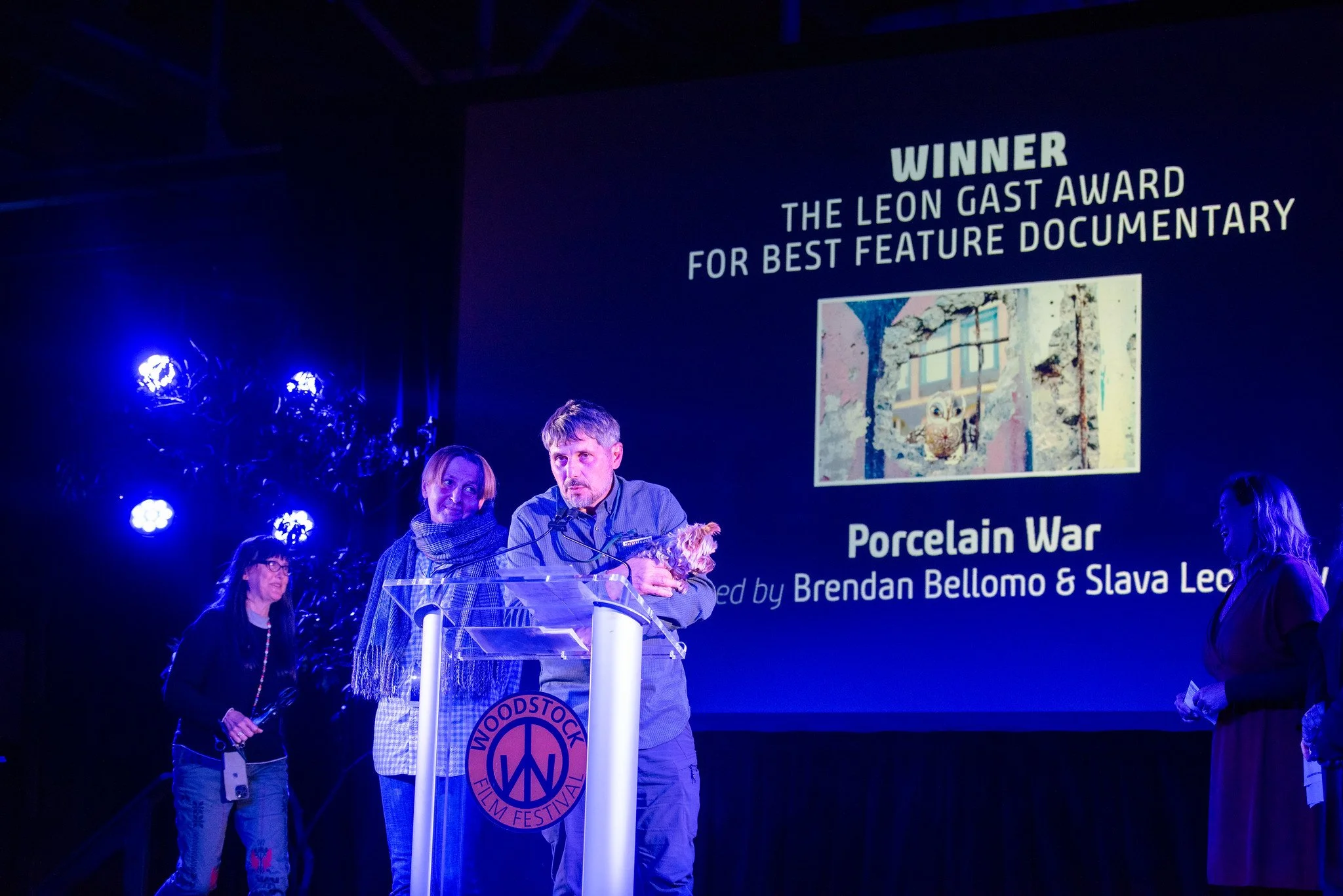 Award ceremony at Woodstock Film Festival with four people on stage, one man holding a small dog, and a large screen behind displaying recognition for the documentary 'Porcelain War', with bright blue lighting.
