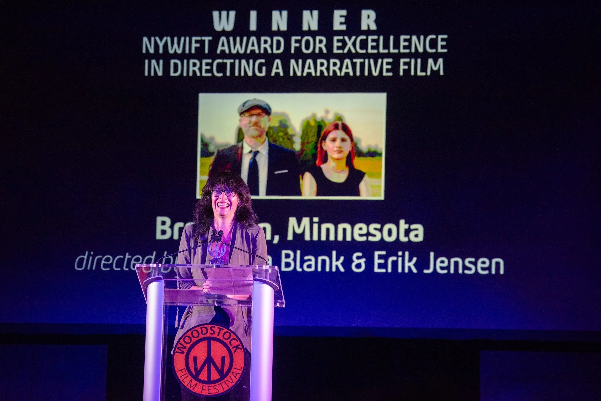 A woman standing behind a clear podium with a Woodstock Film Festival logo, smiling and speaking at an award ceremony on a stage with a large screen behind her. The screen displays text that reads "WINNER NYWIFF AWARD FOR EXCELLENCE IN DIRECTING A NARRATIVE FILM" and features a photo of a man and a woman, along with the text "Brainerd, Minnesota directed by Erik Jensen & Karen Blank."