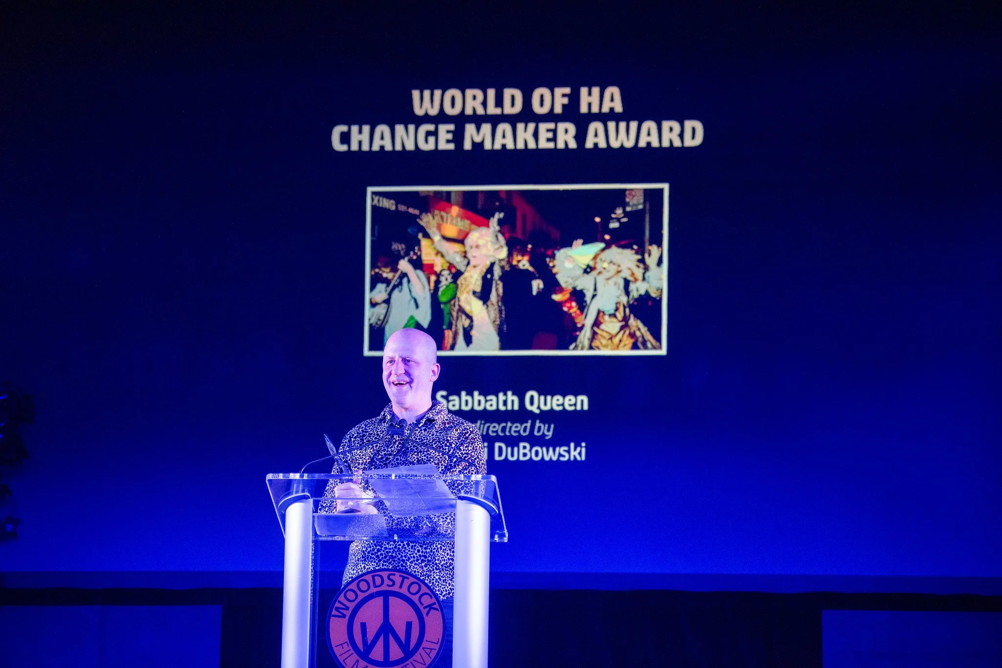 Man speaking at a podium with a large screen behind him displaying the title ‘World of Ha Change Maker Award’ and a picture of people in costume. The event is at Woodstock Film Festival.