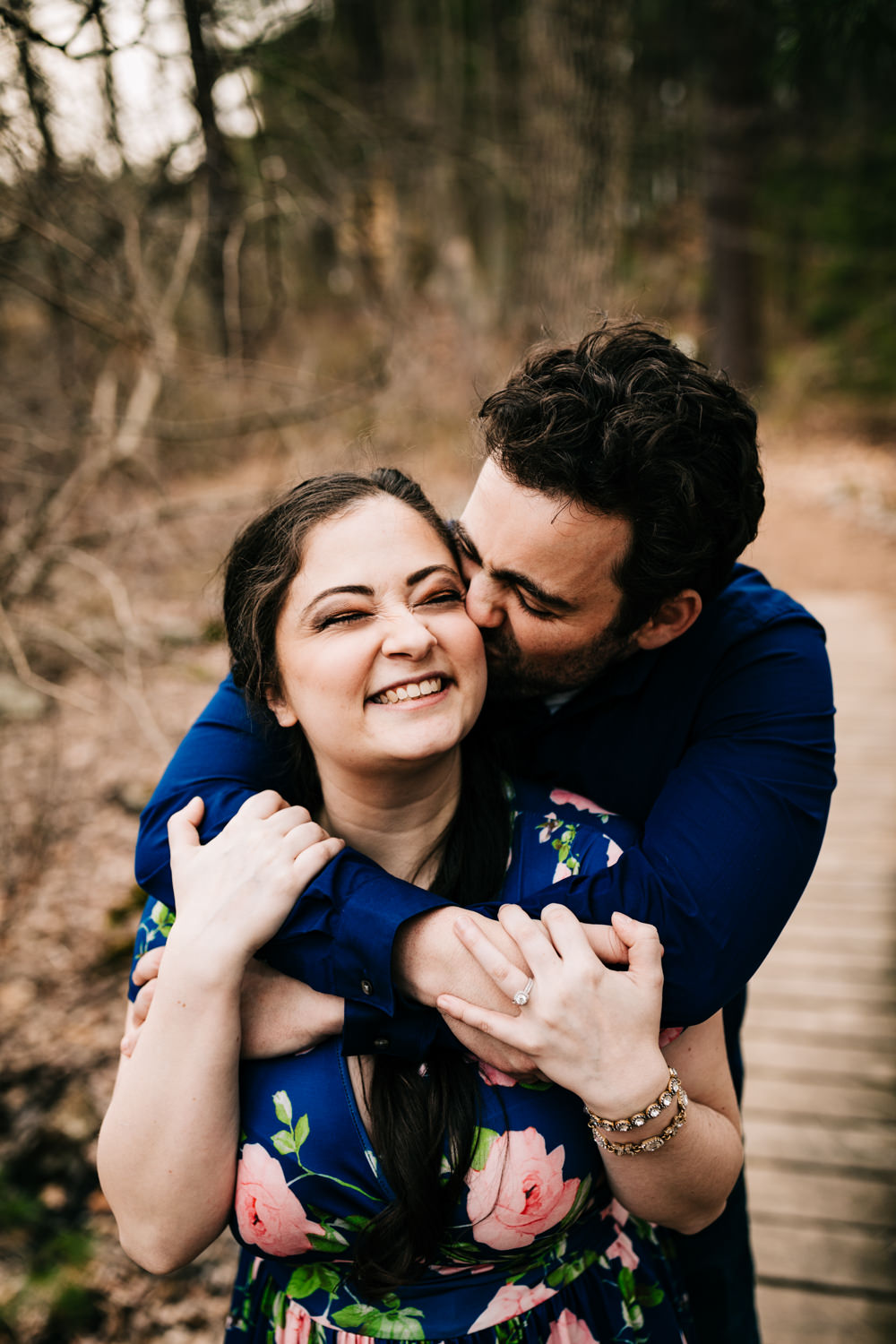 Husband hugging wife from behind wearing blue floral dress in woods of Massachusetts