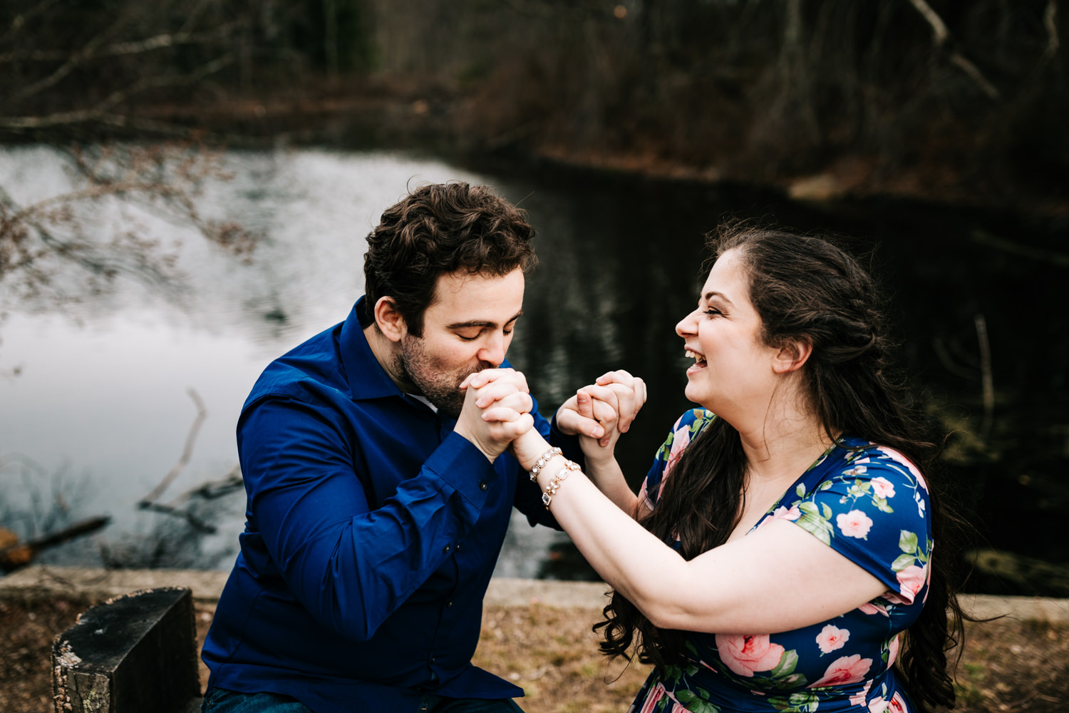 Husband kissing wife's hand wearing blue floral maxi dress near lake in Massachusetts