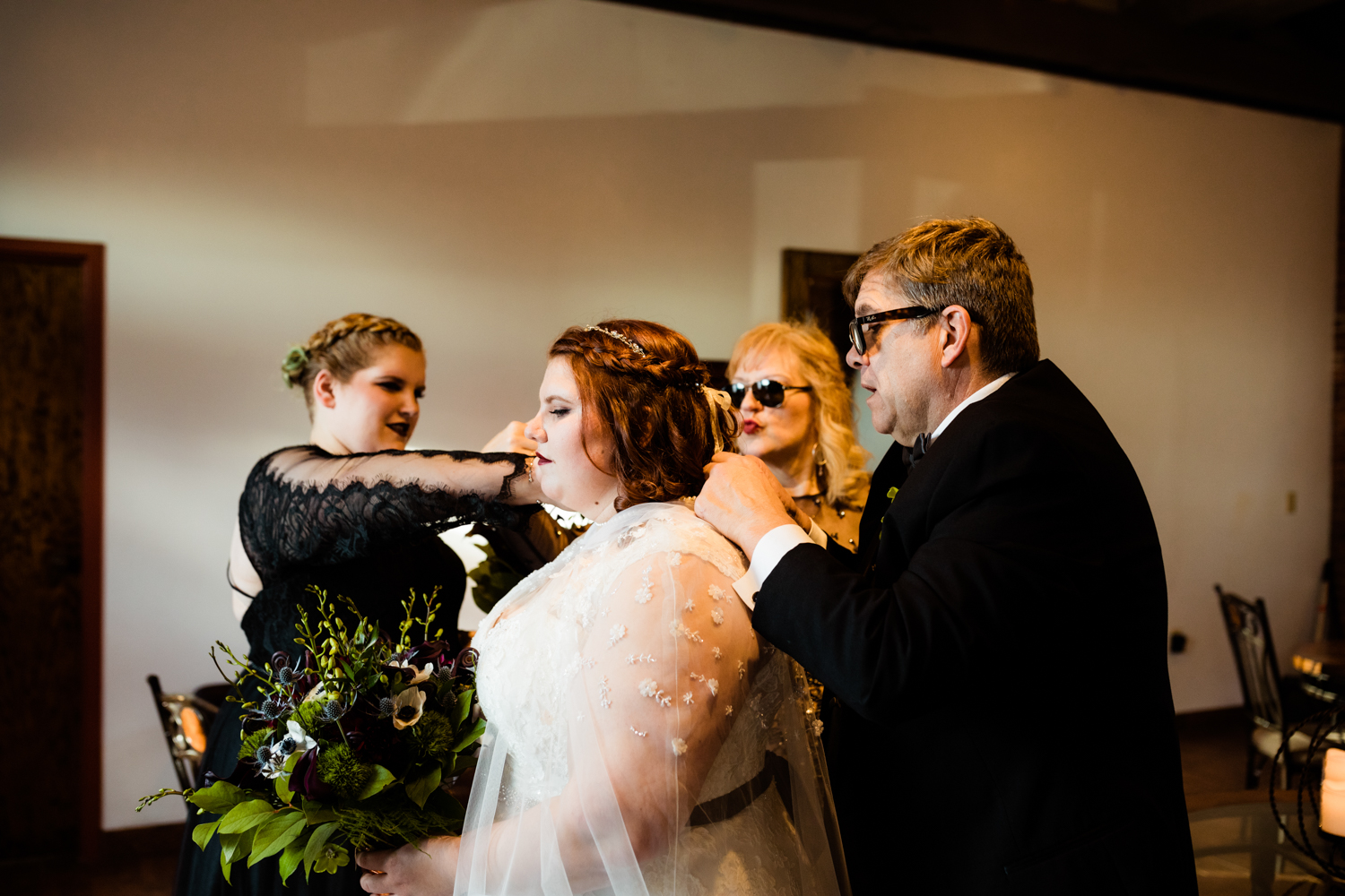 Cape-wearing bride's family helping put on pearl necklace on wedding day