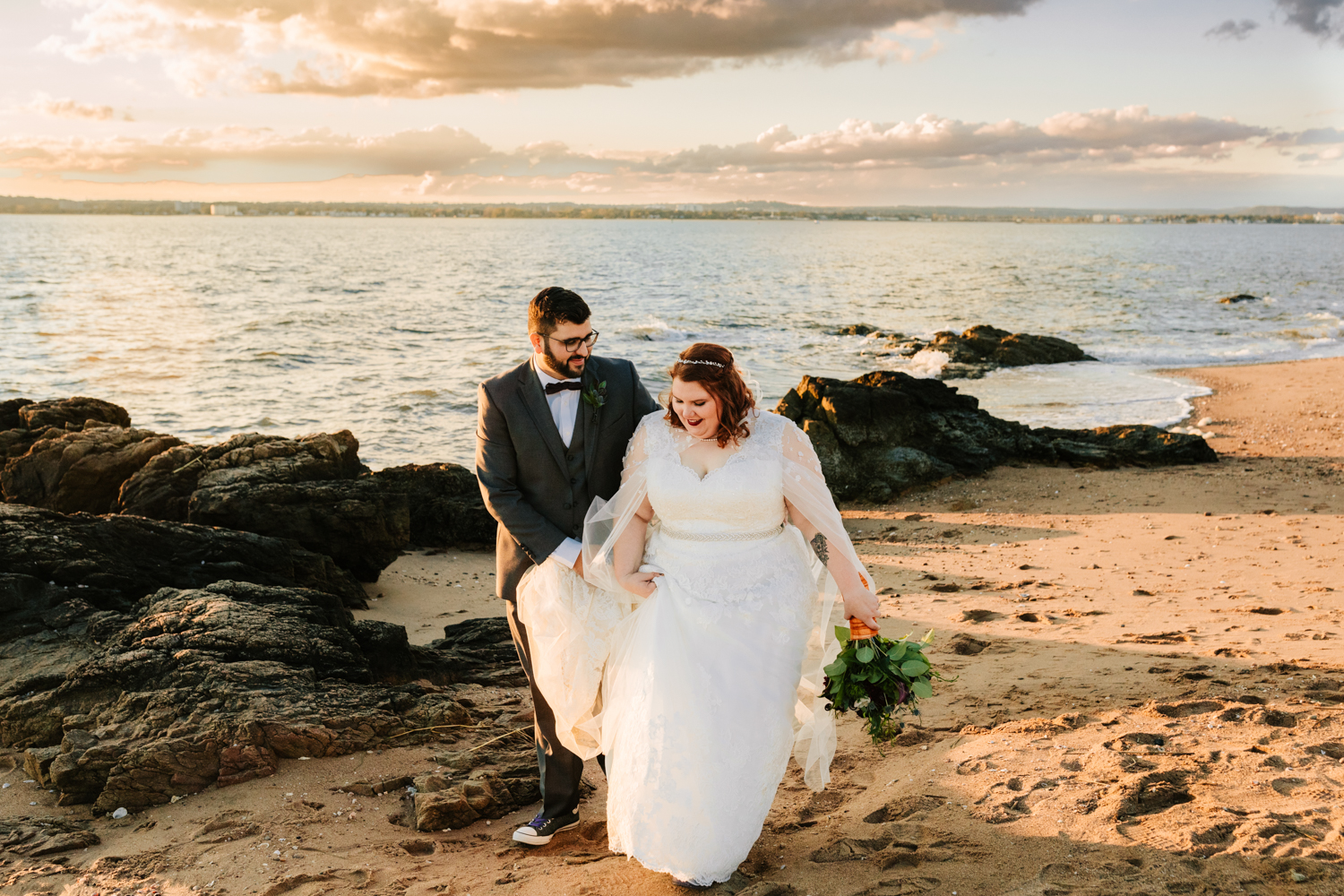 Caped bride and groom walking along beach at sunset