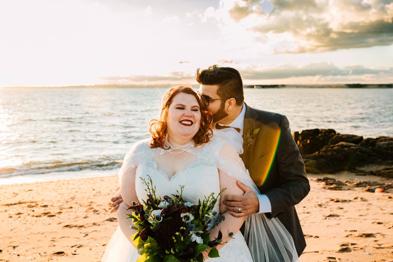 Red haired bride holding bouquet kissed by groom on beach