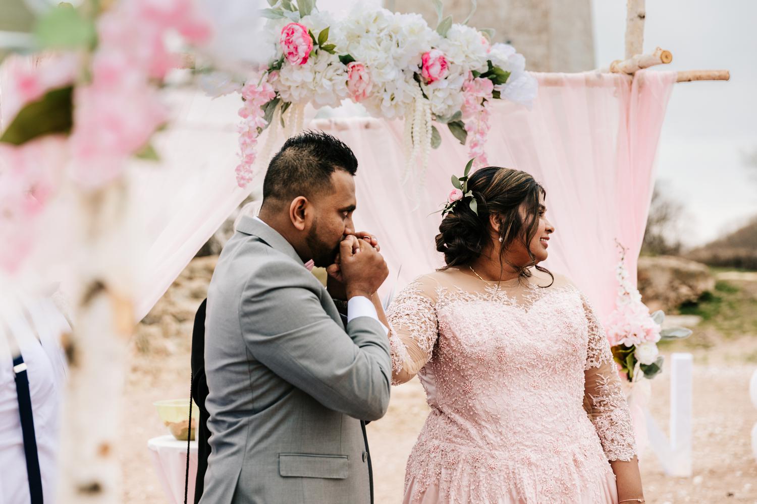 Groom kidding bride's hand during wedding ceremony on beach surrounded by pink and white flowers