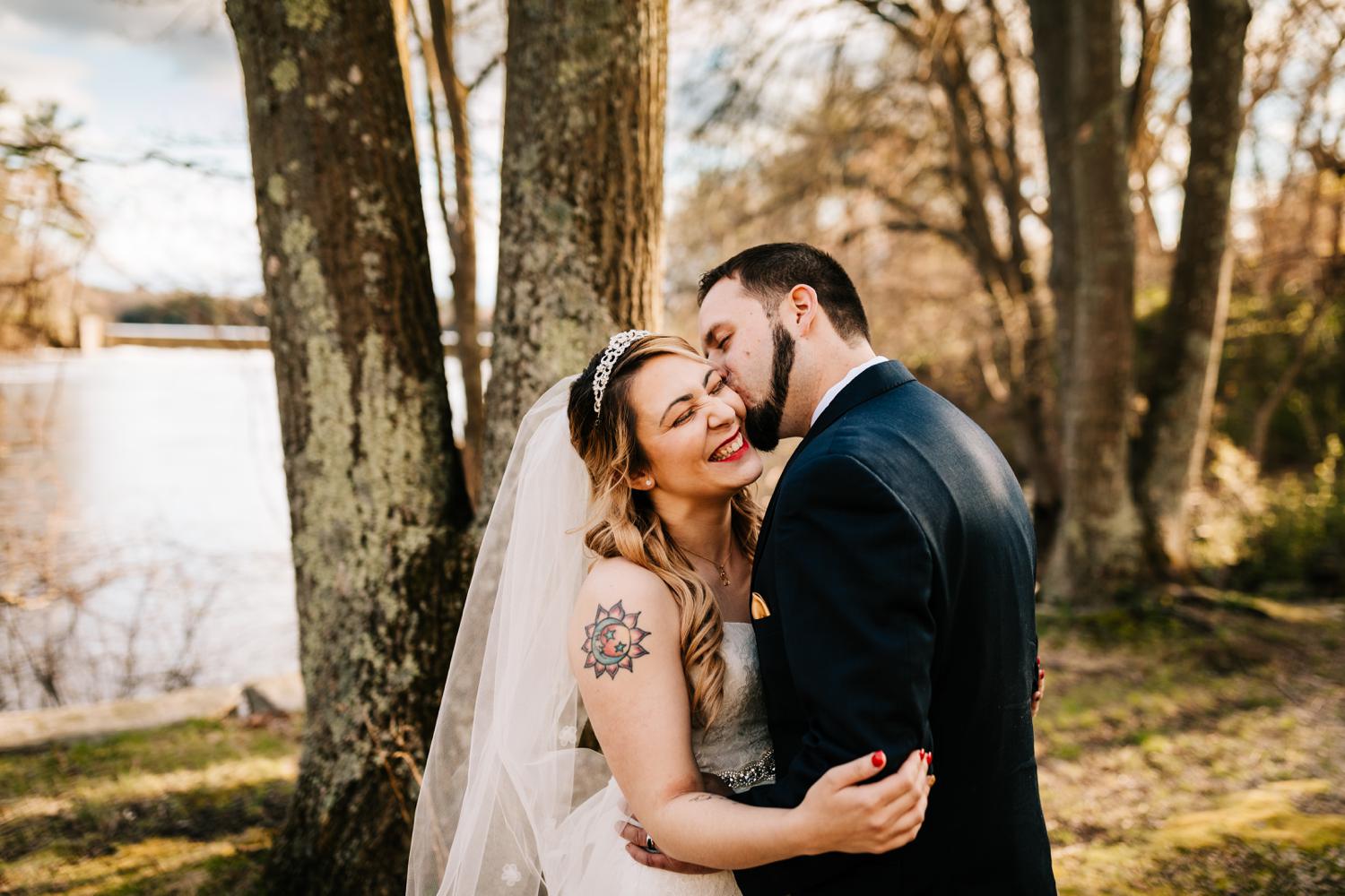 Groom kissing tattooed bride in woods at golden hour