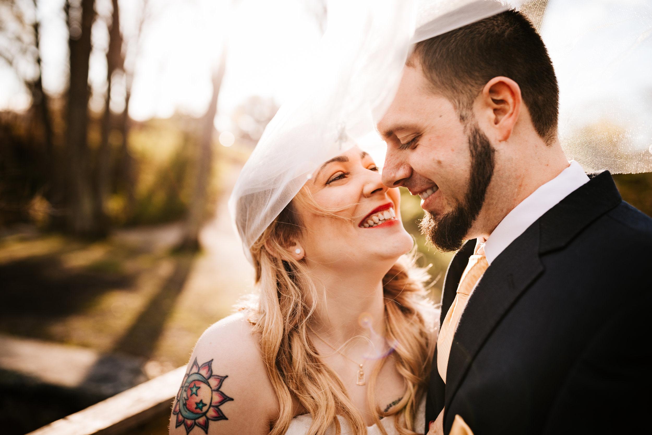 Smiling tattooed bride and groom under veil at golden hour