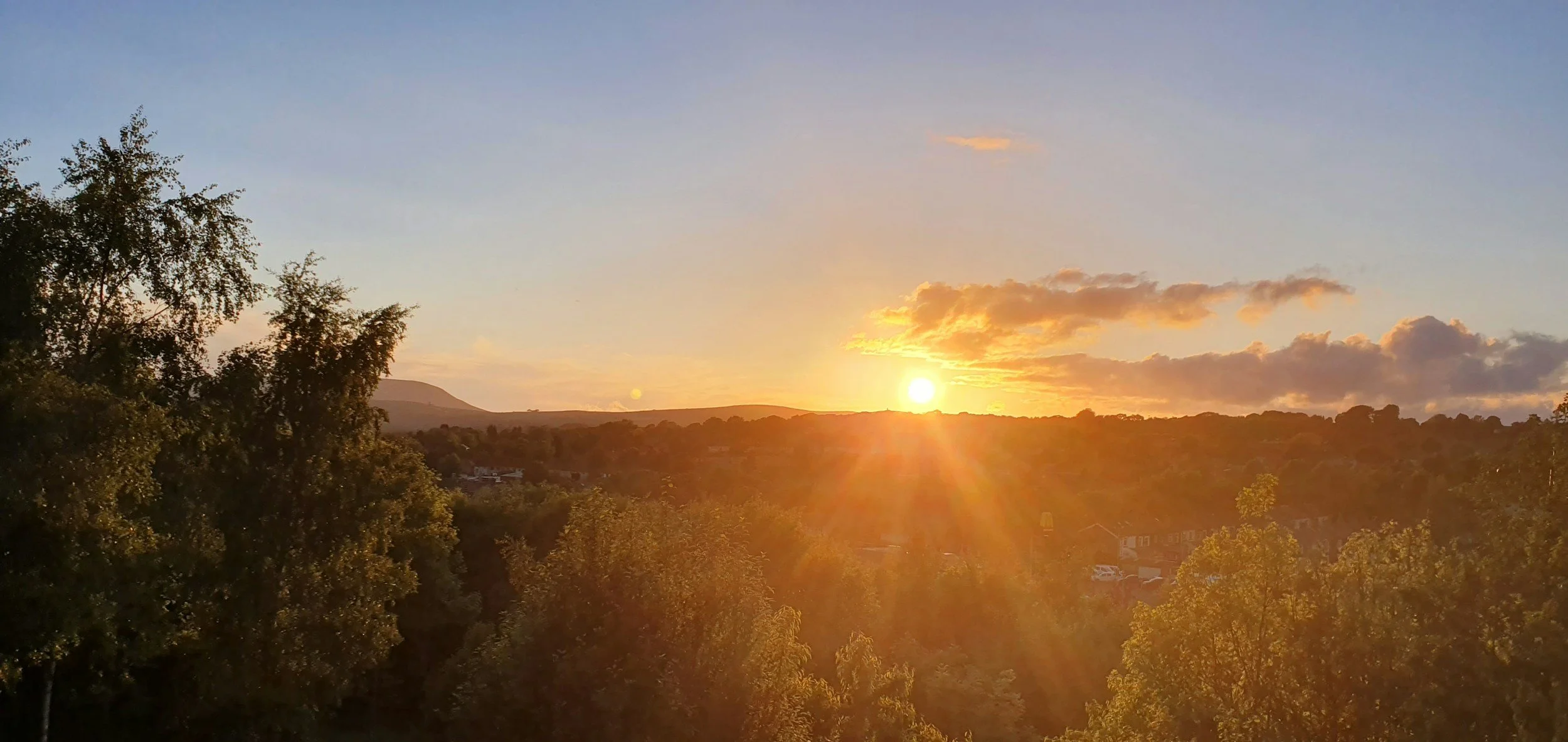 Sunset over a landscape with trees in the foreground and hills in the background, partly cloudy sky with the sun near the horizon.