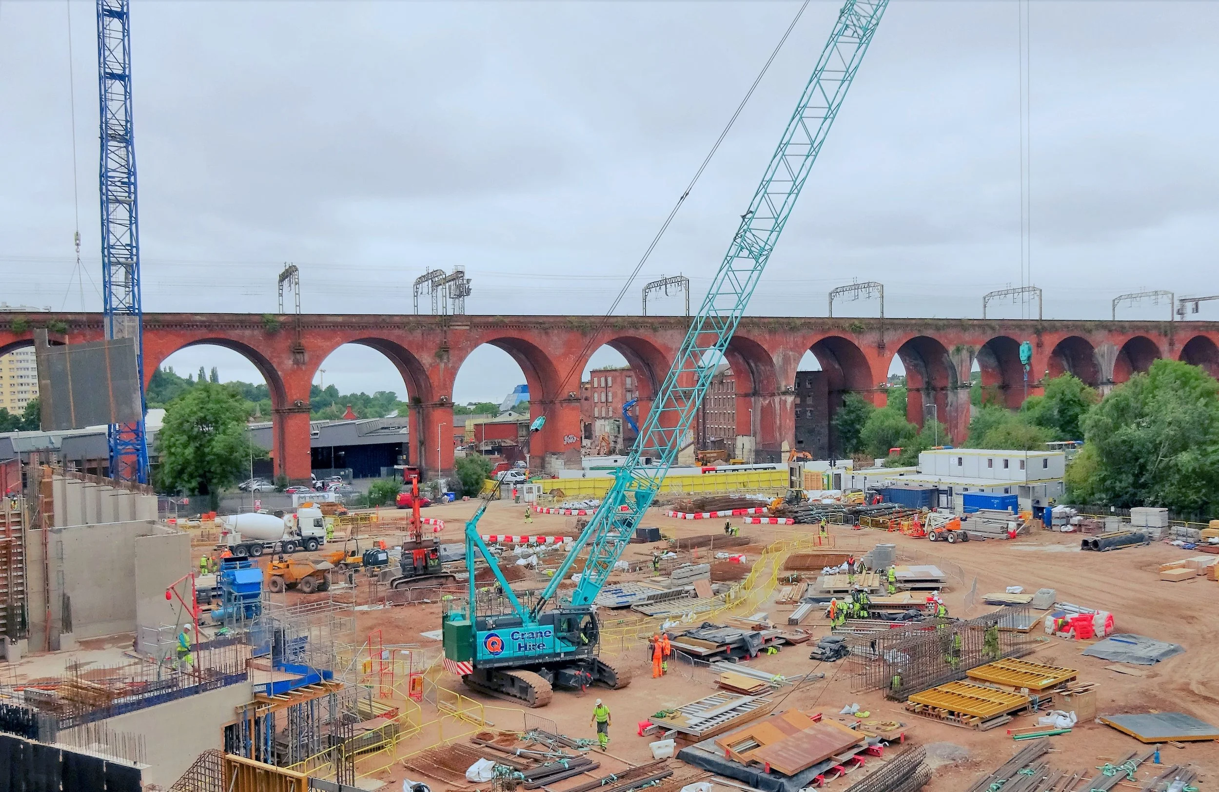 A large construction site with a blue crane, workers, and building materials. In the background, a brick arch bridge with multiple arches and overhead train tracks is visible.