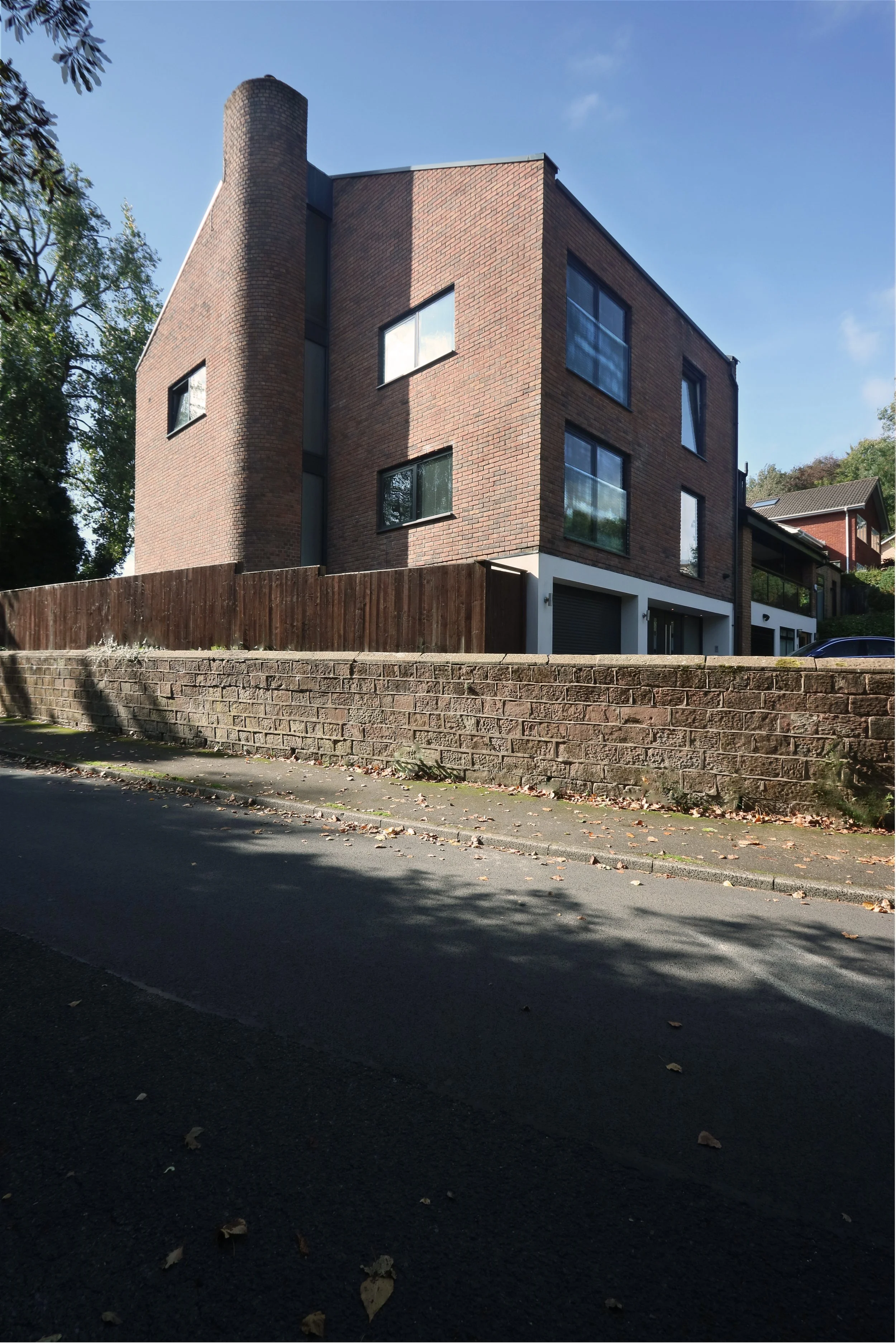 A modern multi-story brick building with large windows and a cylindrical tower on the corner, situated behind a brick and wooden fence, on a sunny day with blue sky and trees in the background.