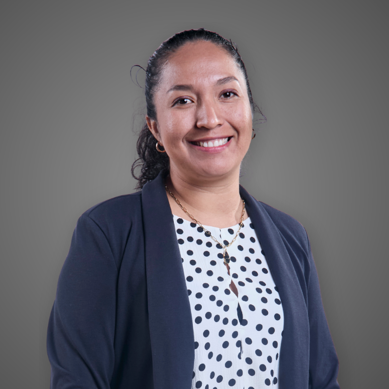 A woman with dark hair tied back, wearing a light blue button-up shirt with a logo on the chest, smiling with her arms crossed, in an indoor setting with bright, blurred lights