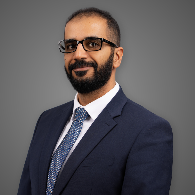 A man with glasses, a beard, and a mustache smiling, wearing a navy blazer and a white shirt, posing in a modern indoor space with bright lighting and blurred background.